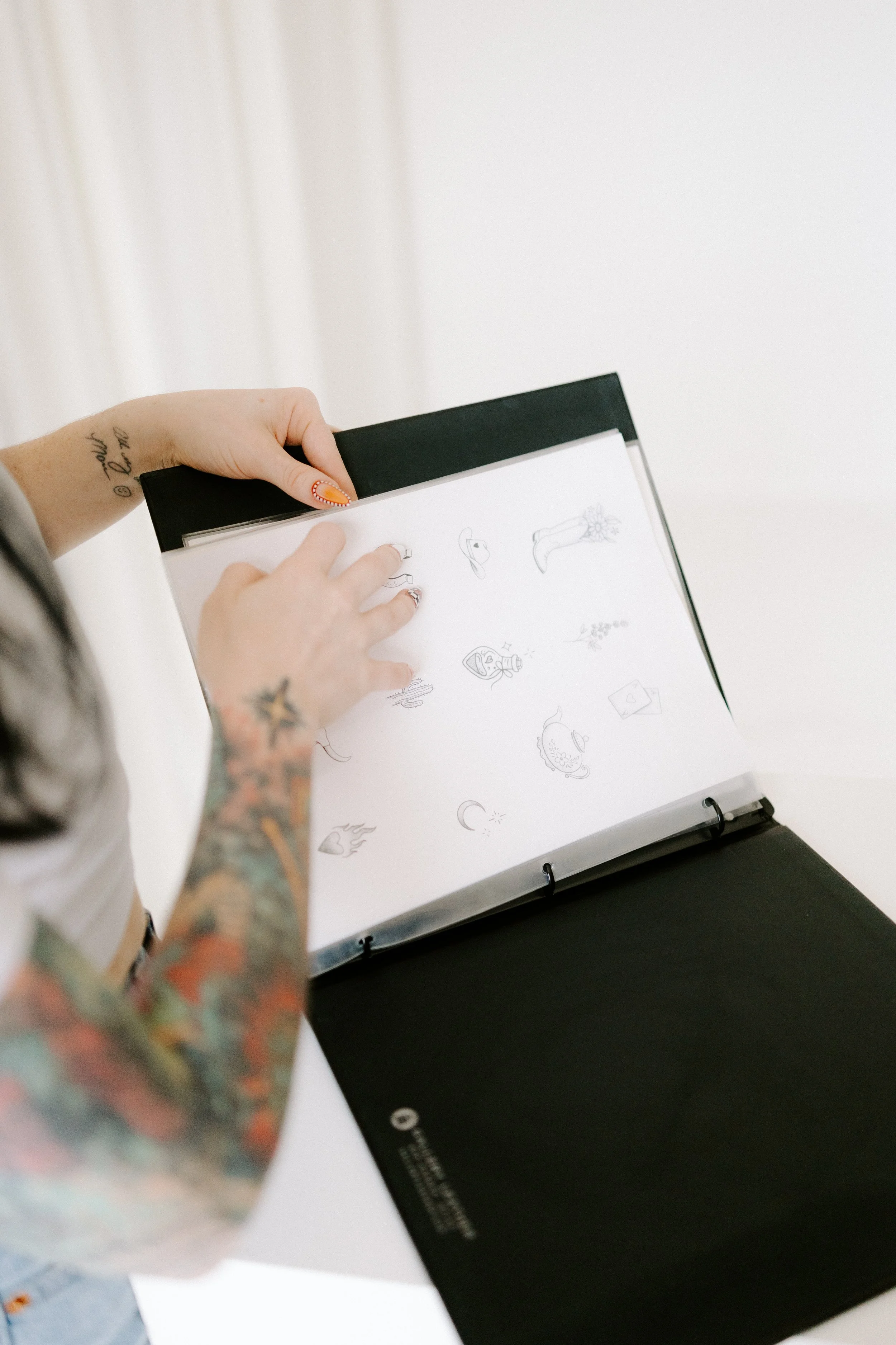 A person with tattoos on their arms is holding a black binder and flipping through pages that have black and white sketches of various items, such as a hat, a bottle, a flower, and a mask.