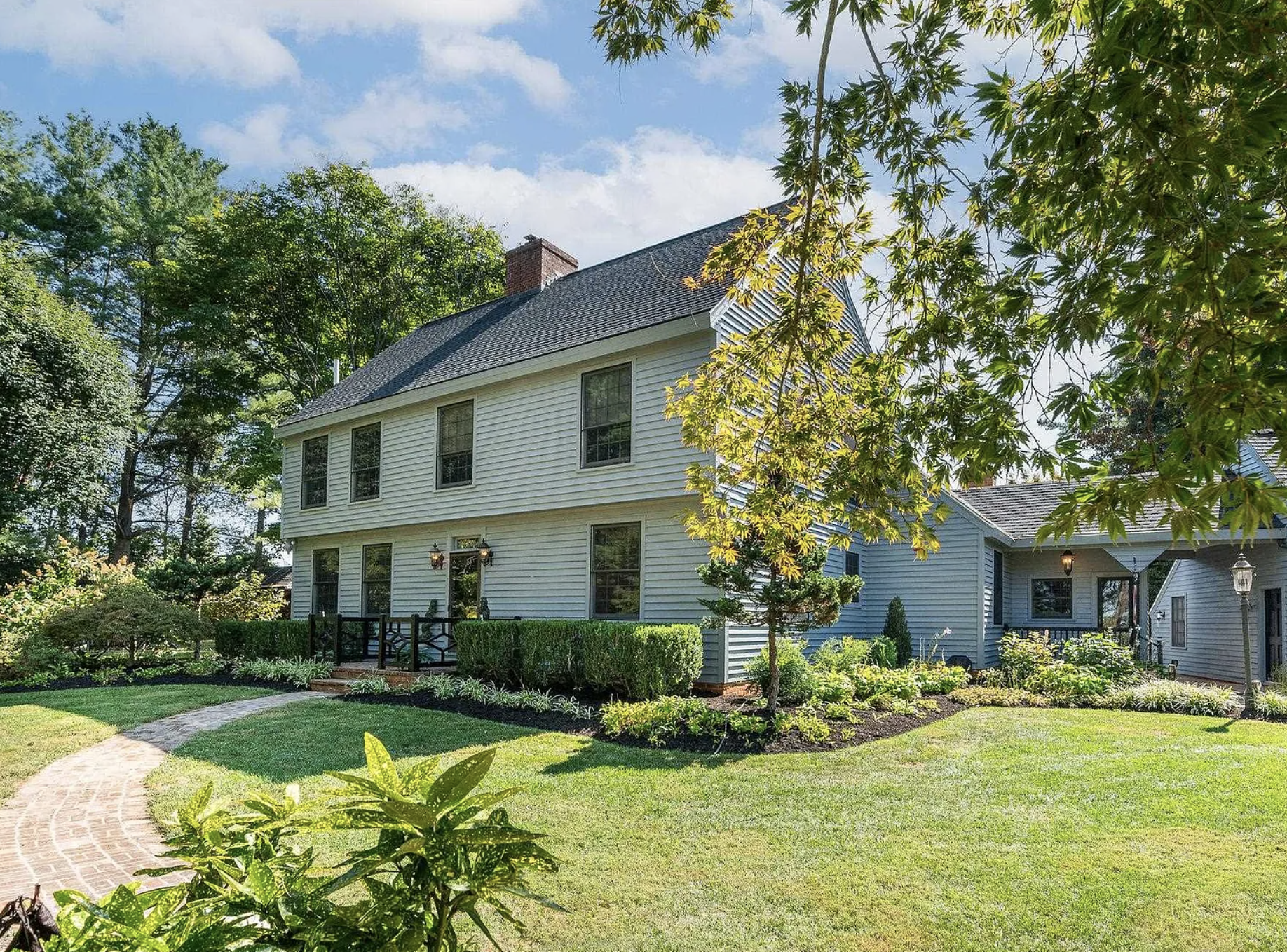 A white two-story house with black lanterns, a small front porch with railing, surrounded by green shrubs, trees, and a well-maintained lawn with a curved brick pathway. 67 Pen y Bry Ln. Fishervile, VA.