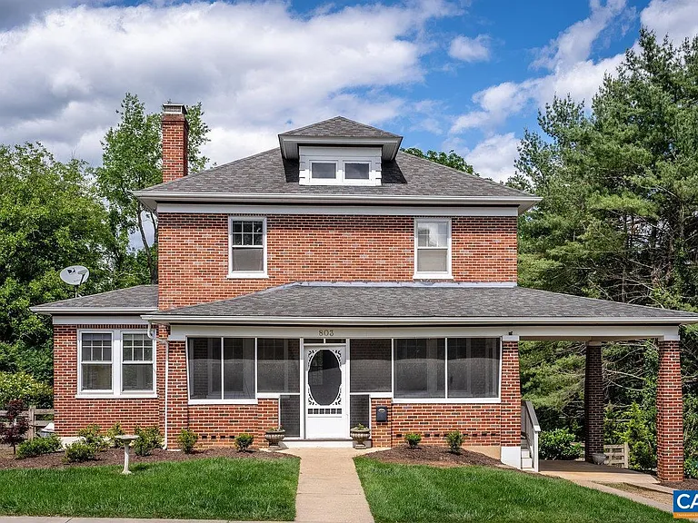 A two-story red brick house with a front porch and gray shingled roof, surrounded by green grass and trees.