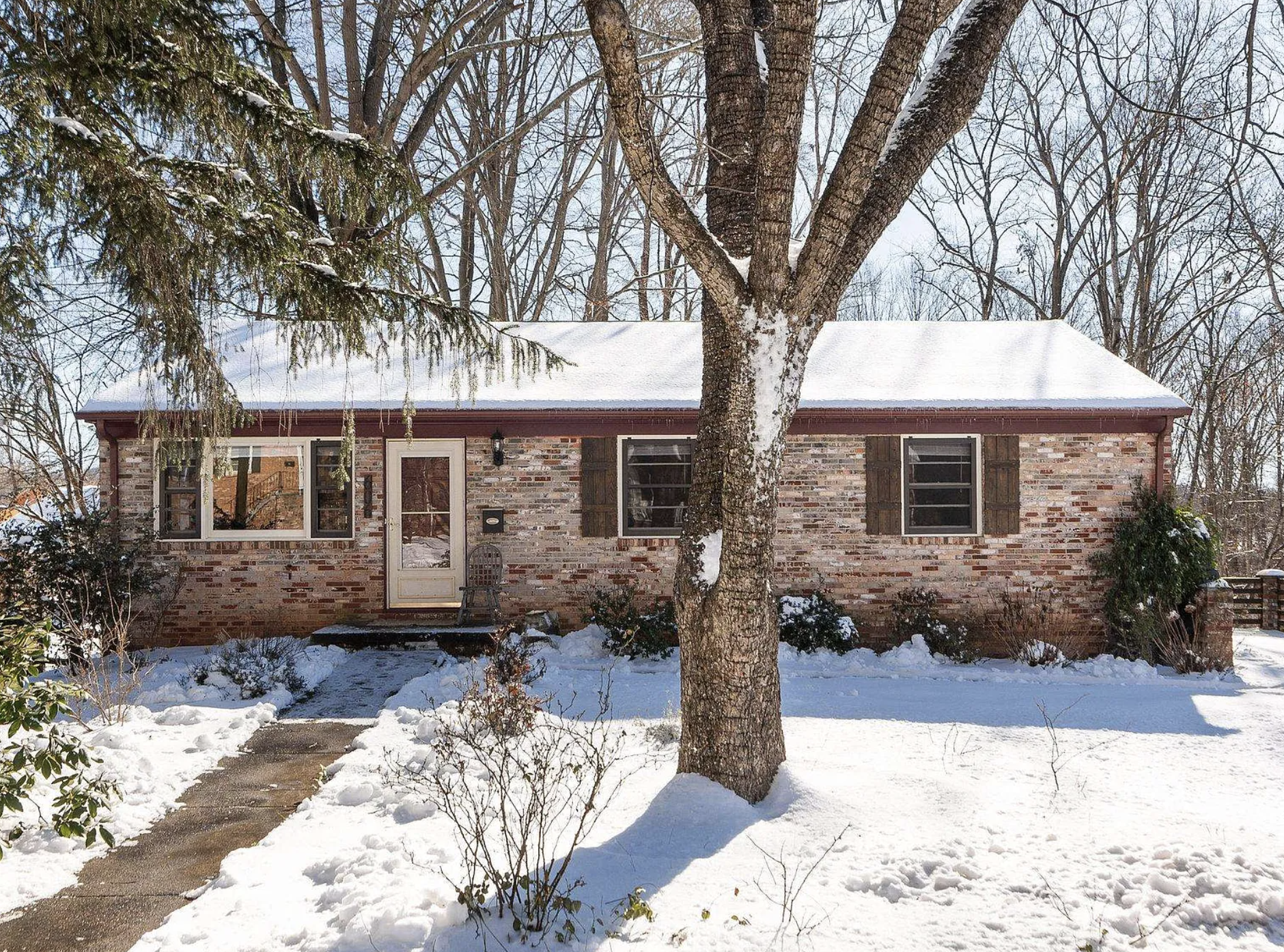 Snow-covered brick house with a large tree in front, surrounded by leafless trees, during winter. 606 Moseley Drive, Charlottesville, VA