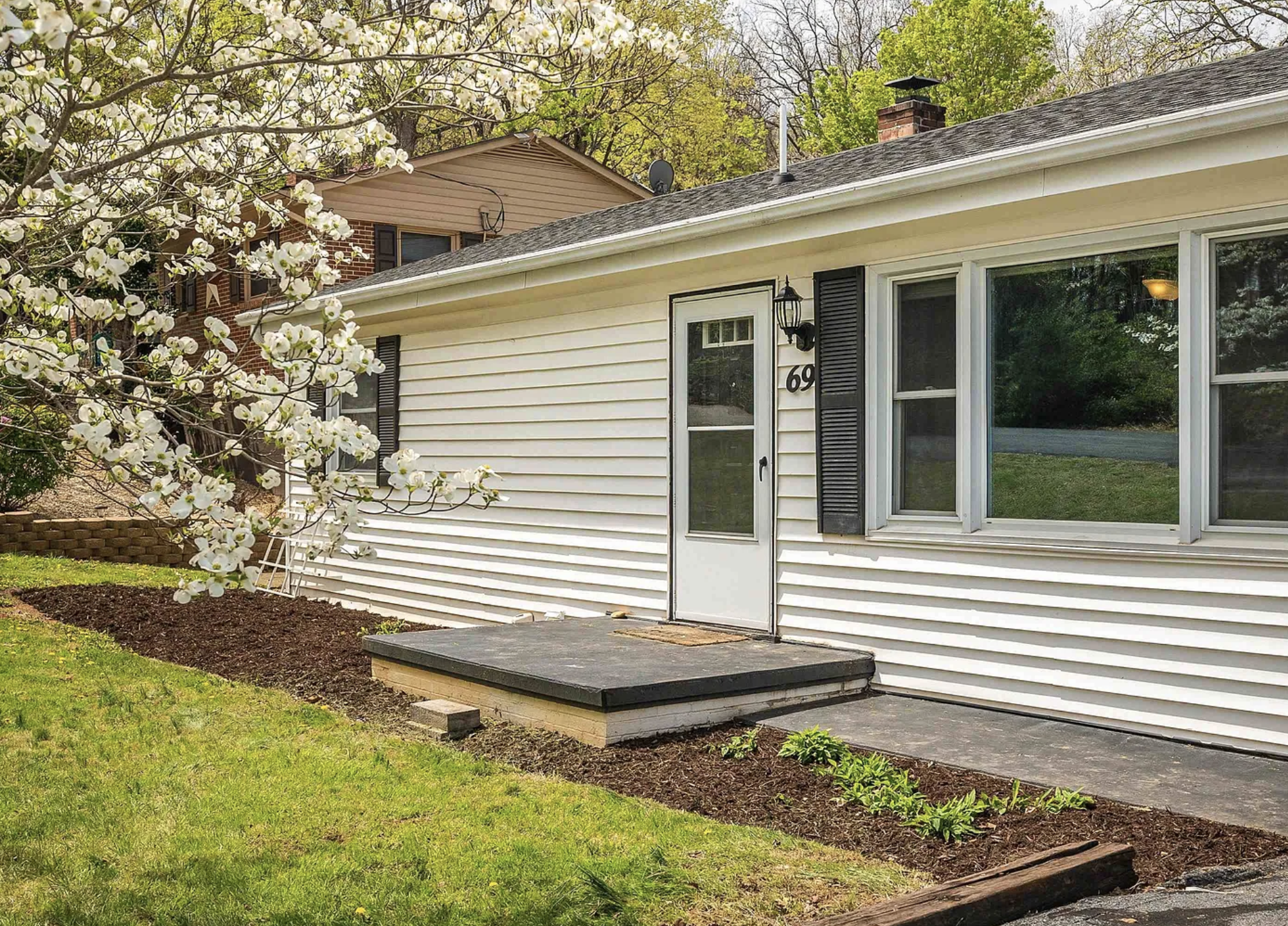 White house with black shutters, front door, and large window, surrounded by green grass, flowering tree, and a garden bed with plants. 69 Jefferson St. Verona, VA.