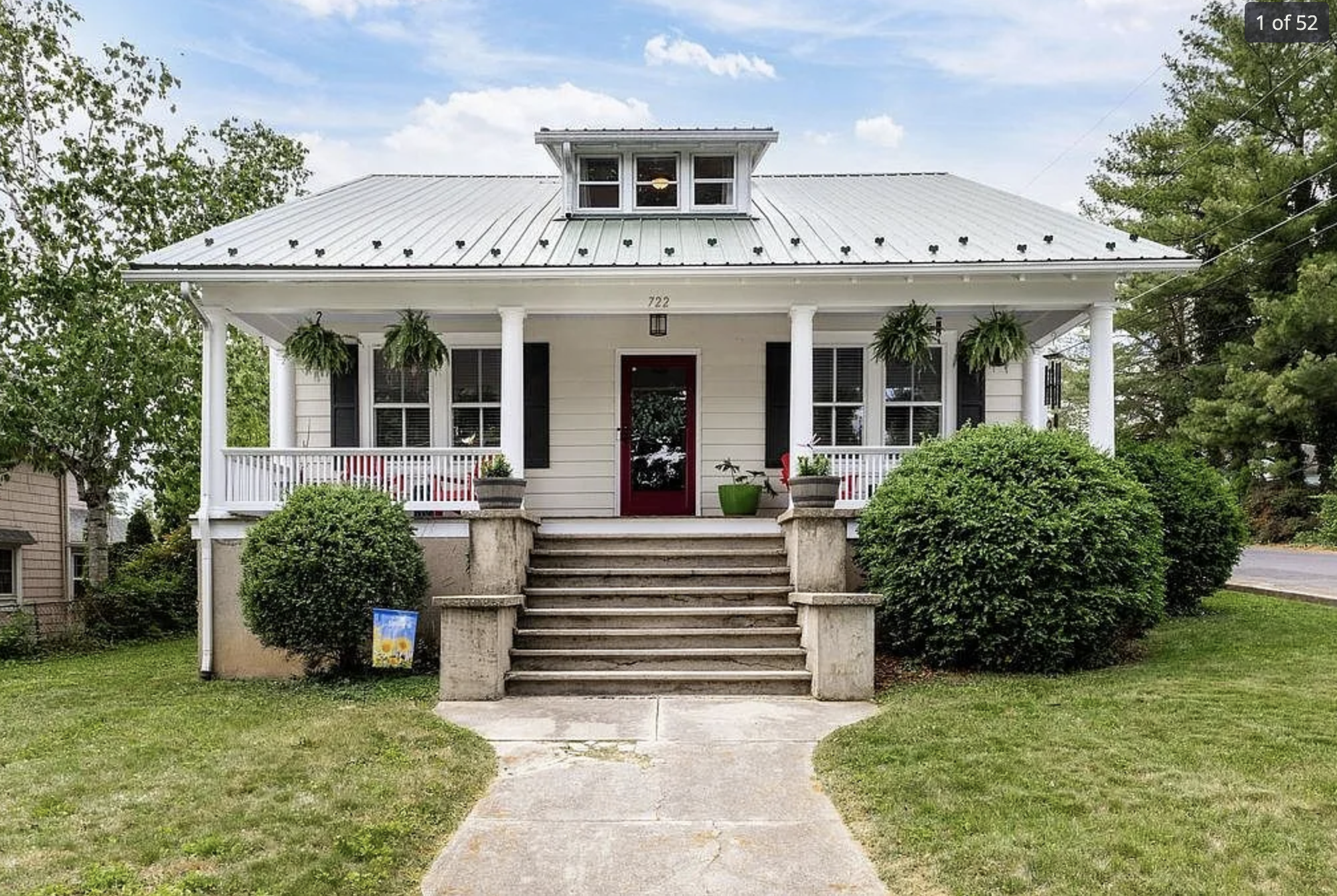 Front view of a white house with a metal roof, front porch with potted plants, hanging ferns, bushes, and a concrete walkway leading to stairs. 722 Alleghany Ave. Staunton, VA.