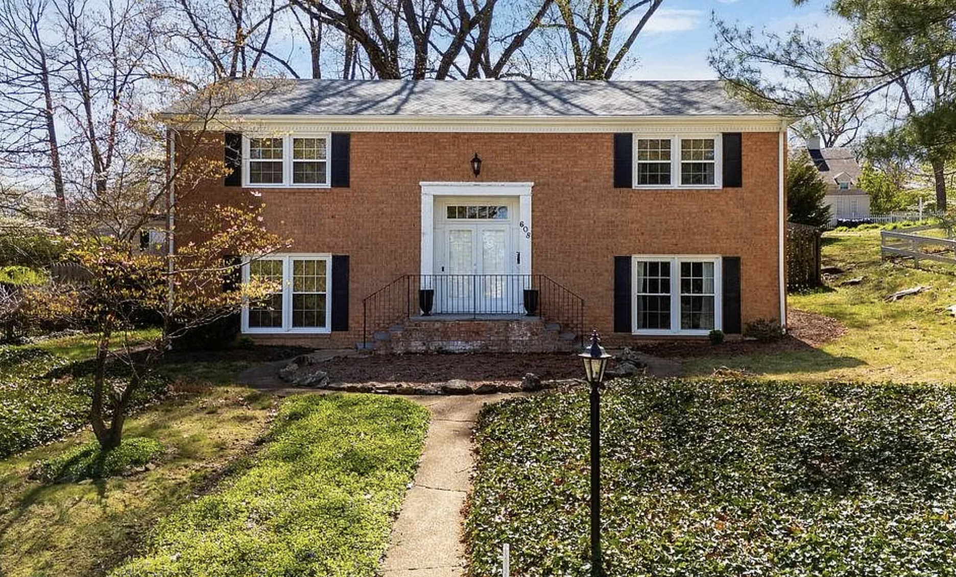 Front view of a two-story brick house with white door, five windows with white frames and black shutters, front stairs, landscaped yard, small trees, a lamp post, and neighboring houses visible in the background. 608 S Poplar Ave, Waynesboro, VA.