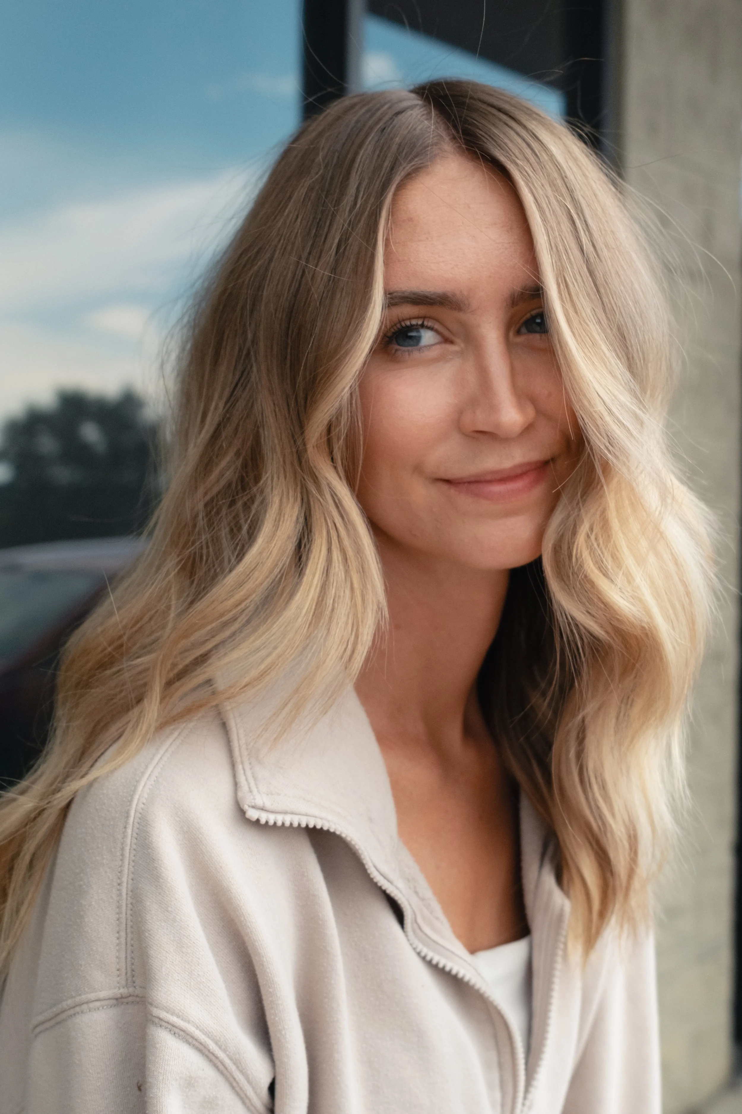 A young woman with long, wavy blonde hair and blue eyes smiling softly in front of a window.