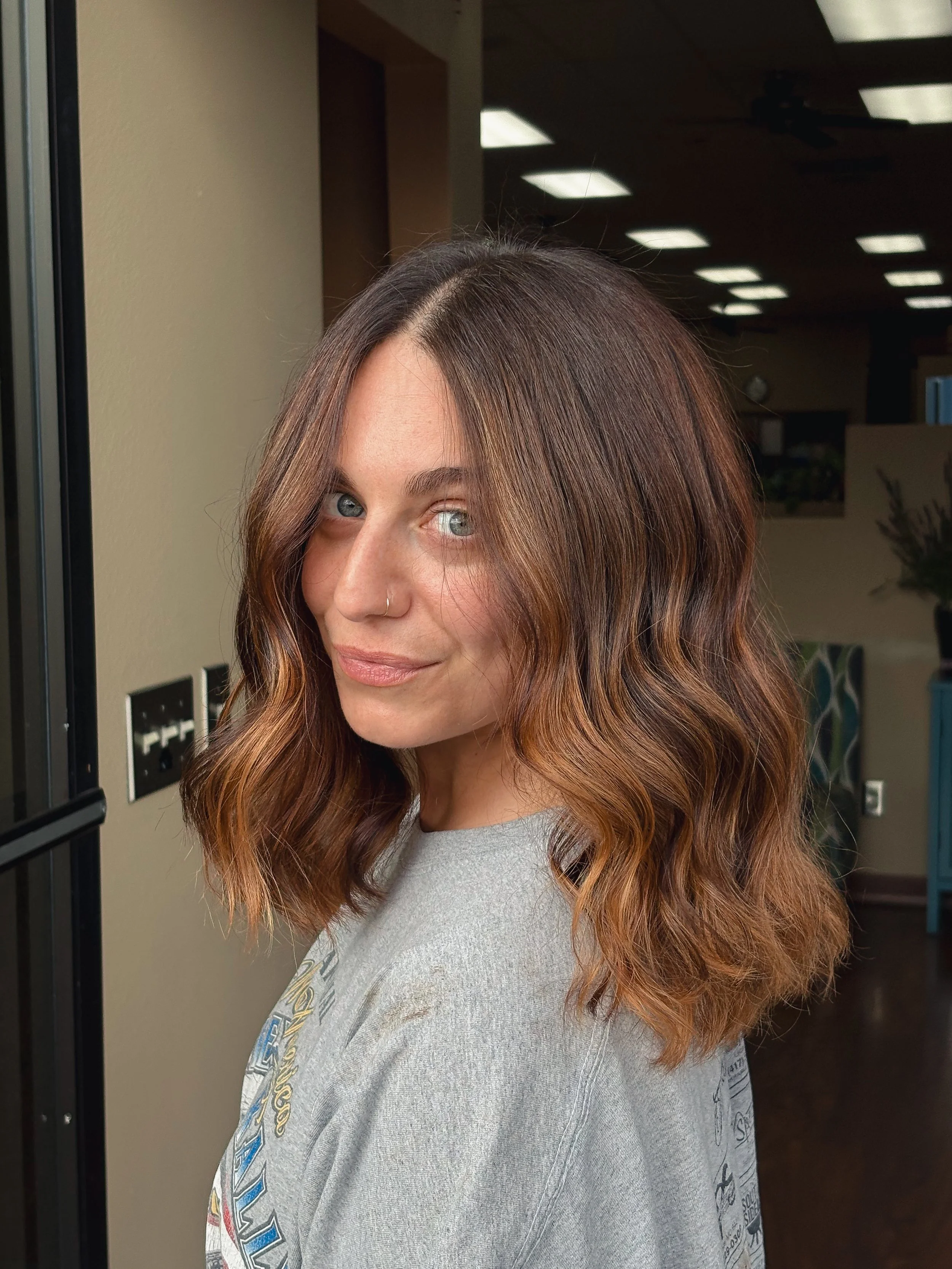 Woman with wavy, medium-length hair with highlights, standing indoors near a glass door, wearing a gray shirt, in an office or salon setting.