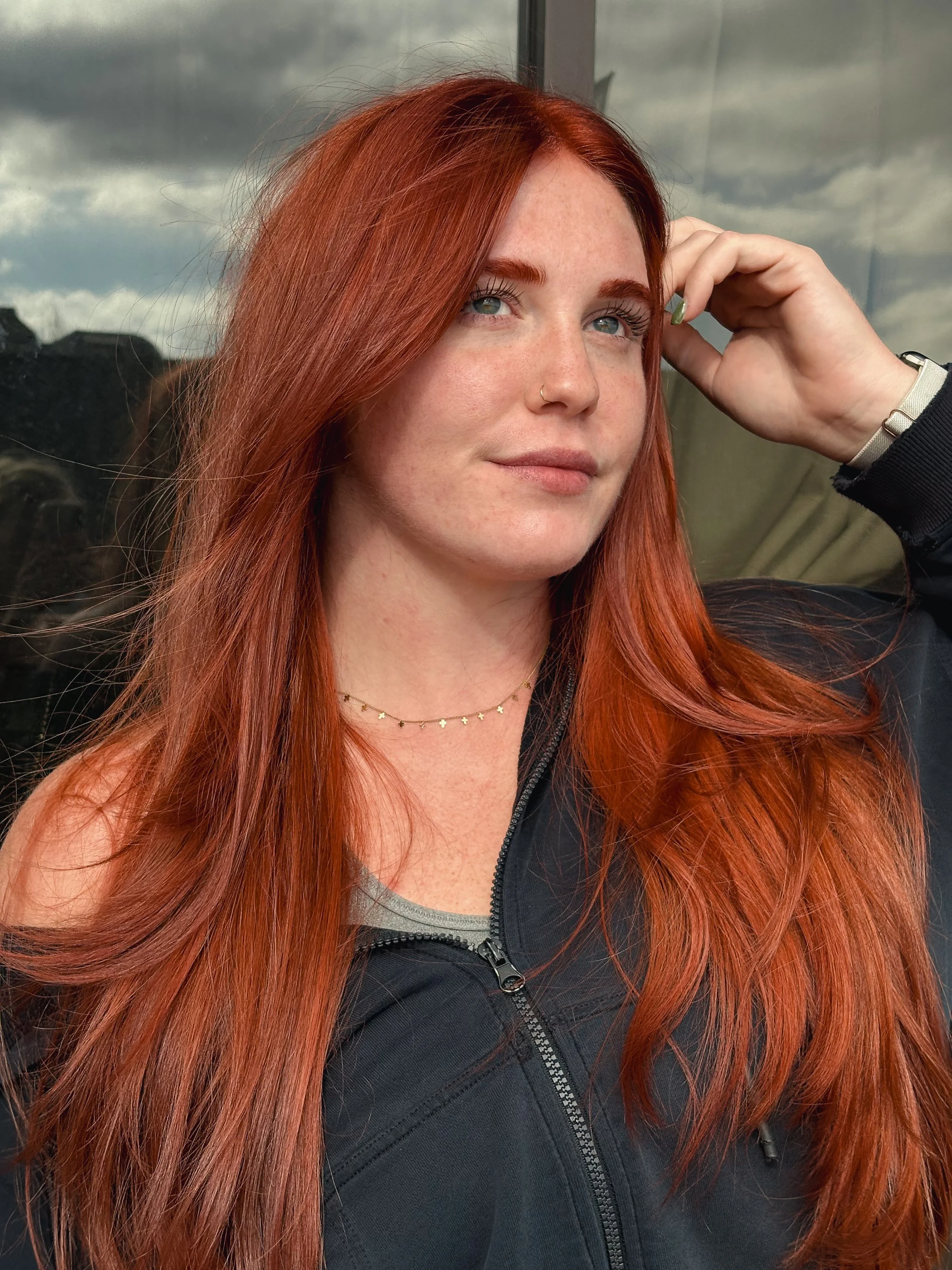 A woman with long red hair, blue eyes, and light skin, wearing a black jacket, a necklace, a nose ring, and a bracelet, standing outdoors against a cloudy sky.
