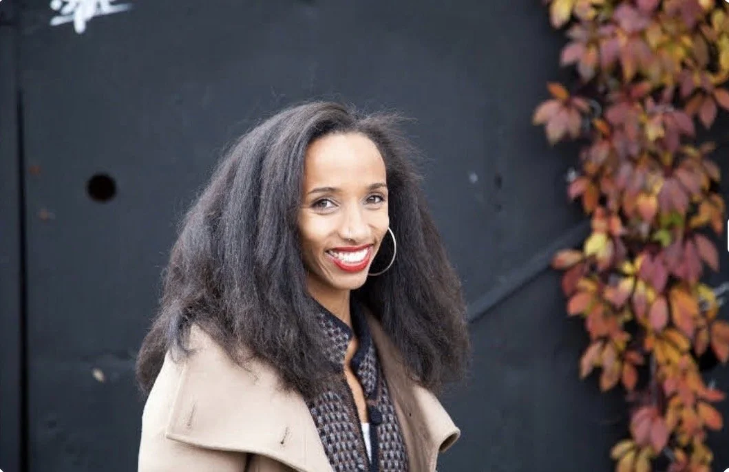 A woman with black, curly hair, red lipstick, and hoop earrings smiling outdoors next to a black wall and autumn-colored leaves.