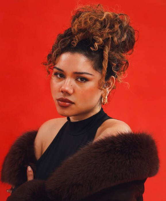 A woman with curly hair styled in an updo, wearing a black sleeveless top and a dark fur coat, posing against a red background.