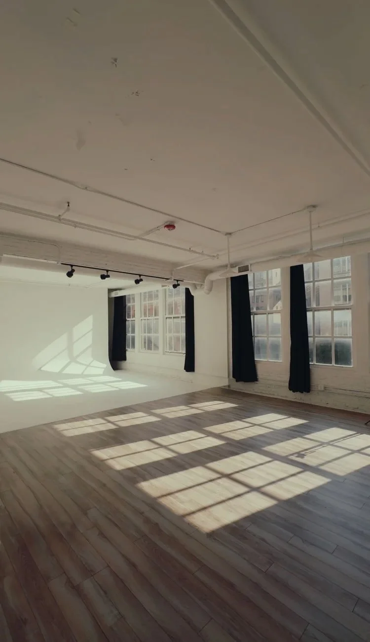 Empty room with wooden floor, large windows, and black curtains, sunlight casting shadows on the floor.