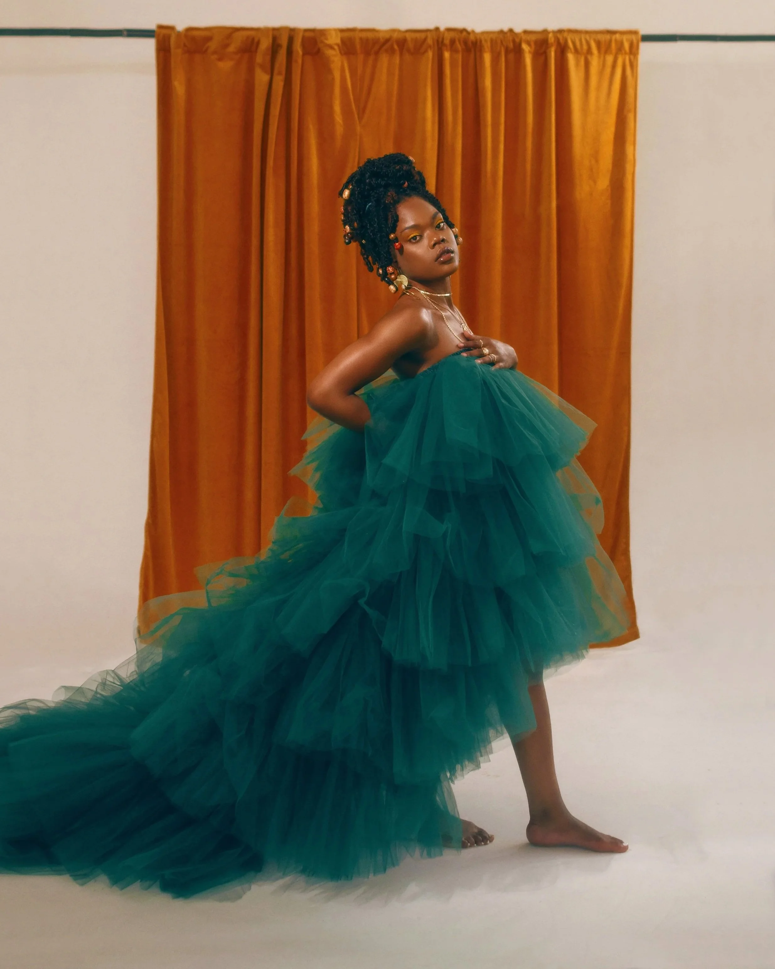A woman with dreadlocks styled in an updo, adorned with beads, wearing a strapless green tulle gown, standing in front of an orange curtain backdrop and white wall, with one hand on her chest and the other on her hip.
