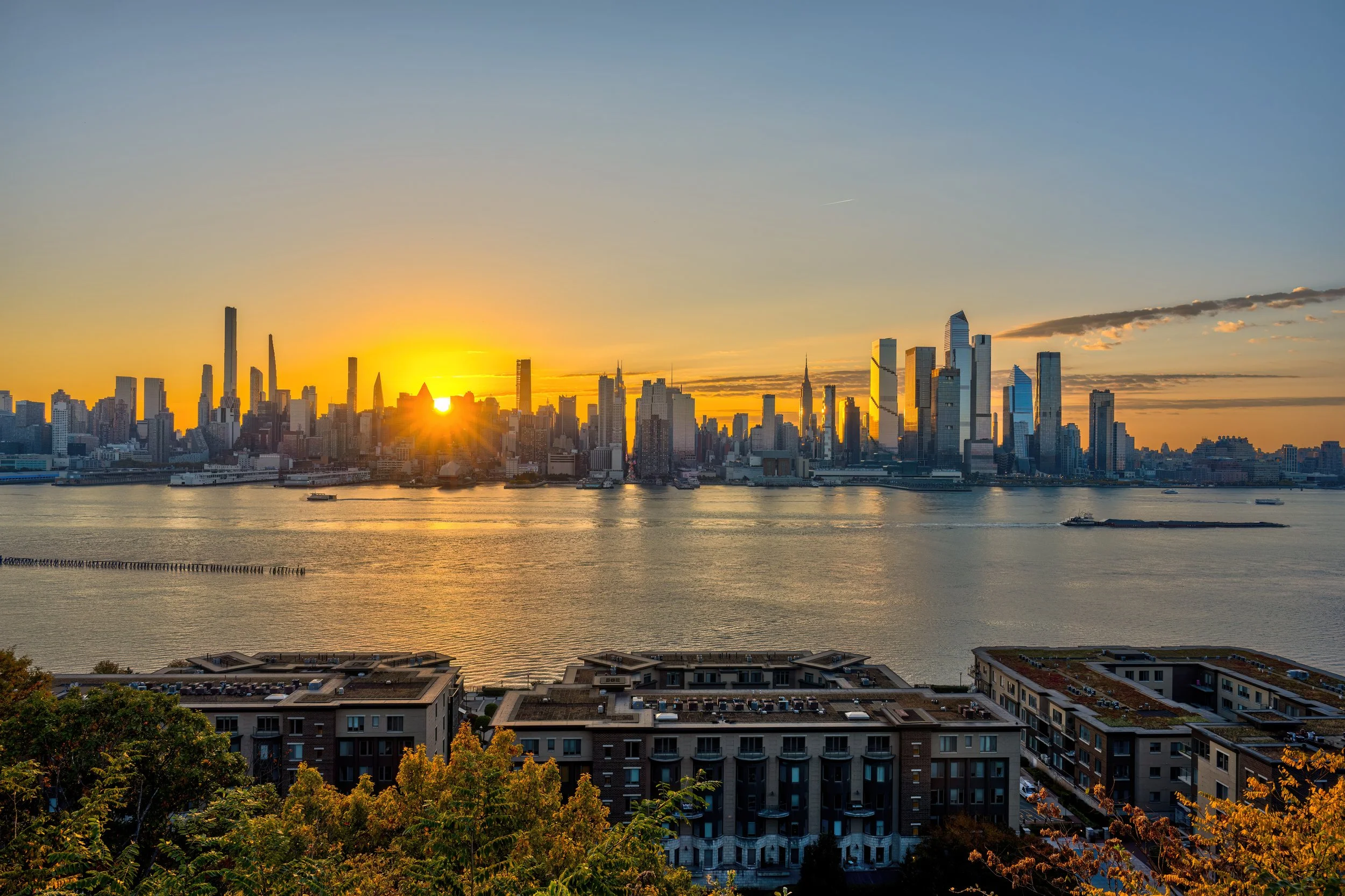 An elevated sunrise view across the Hudson River featuring the Manhattan skyline, with Weehawken residential buildings with green roofs and trees in the foreground.
