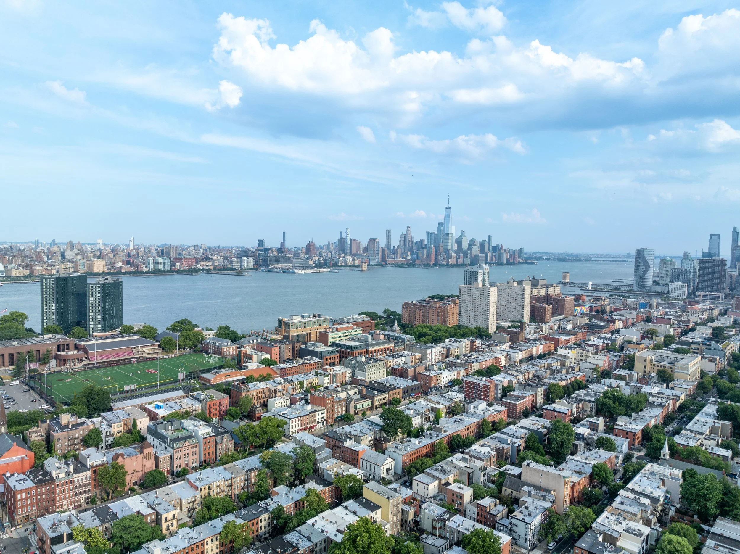 A high-angle cityscape of Hoboken, NJ, looking across the Hudson River toward the One World Trade Center and the Lower Manhattan skyline under a blue sky with light clouds.
