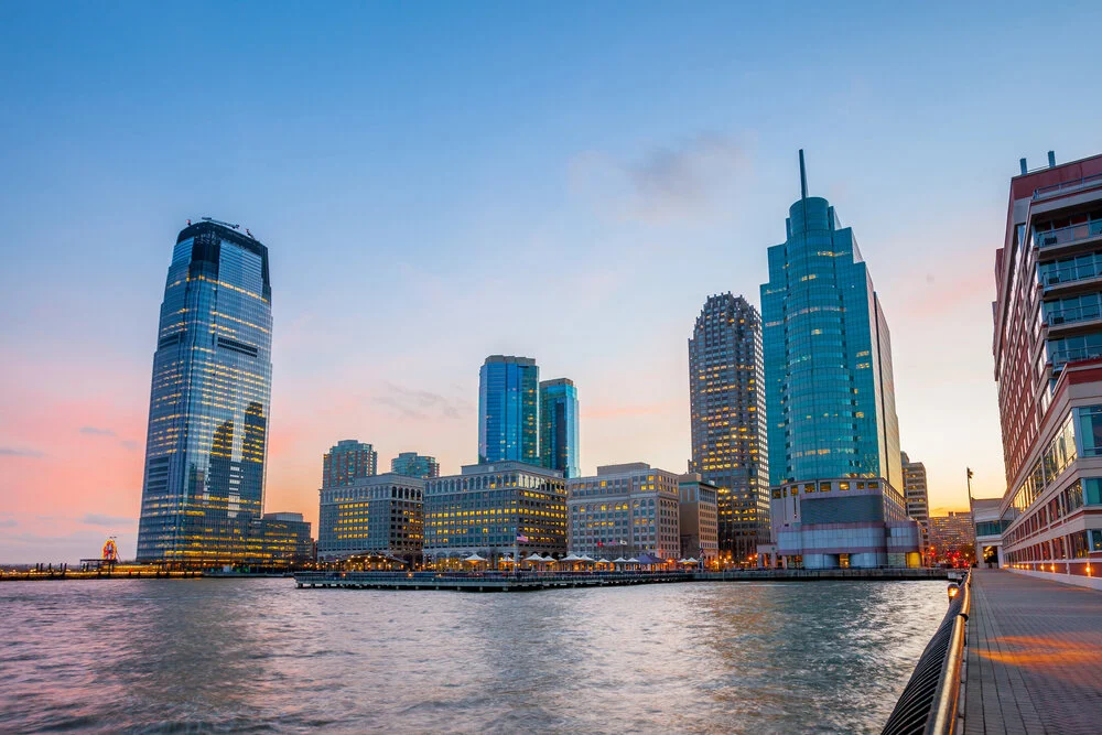 Panoramic view of the Downtown Jersey City waterfront skyline and high-rise towers glowing at dusk along the Hudson River promenade.