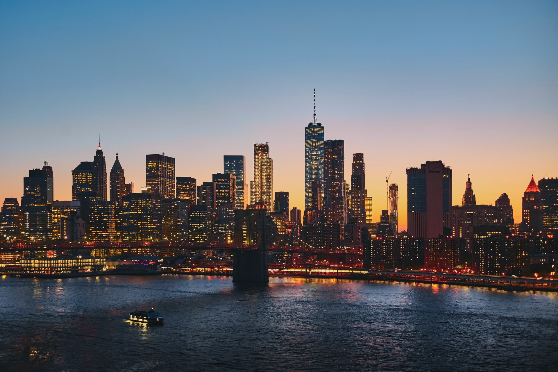 Panoramic view of the glowing city skyline at sunset across the water with an illuminated bridge in the foreground.