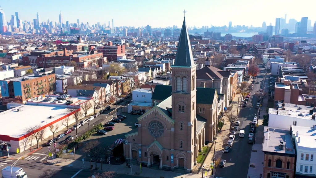 Aerial view of historic St. Lucy's Church on Grove Street surrounded by Downtown Jersey City residential buildings and the Manhattan skyline in the distance.