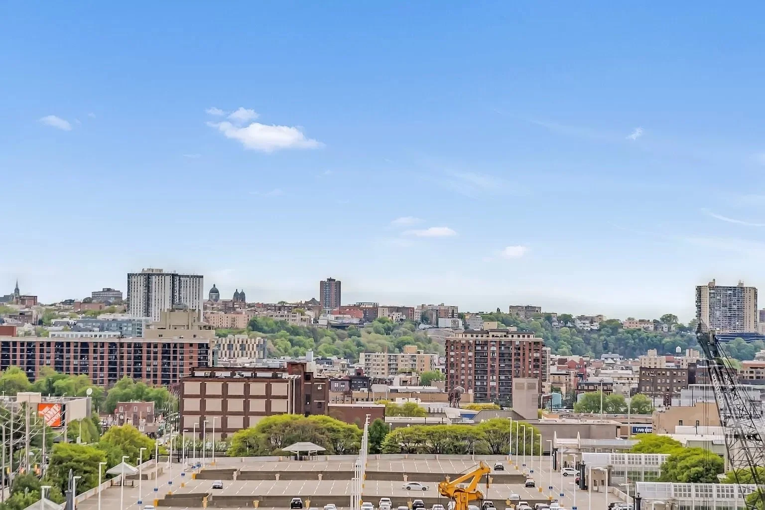 An elevated, expansive view of the Jersey City and Hoboken area landscape, featuring a mix of commercial spaces, large parking areas, and residential mid-rise buildings nestled along the tree-lined hillside.