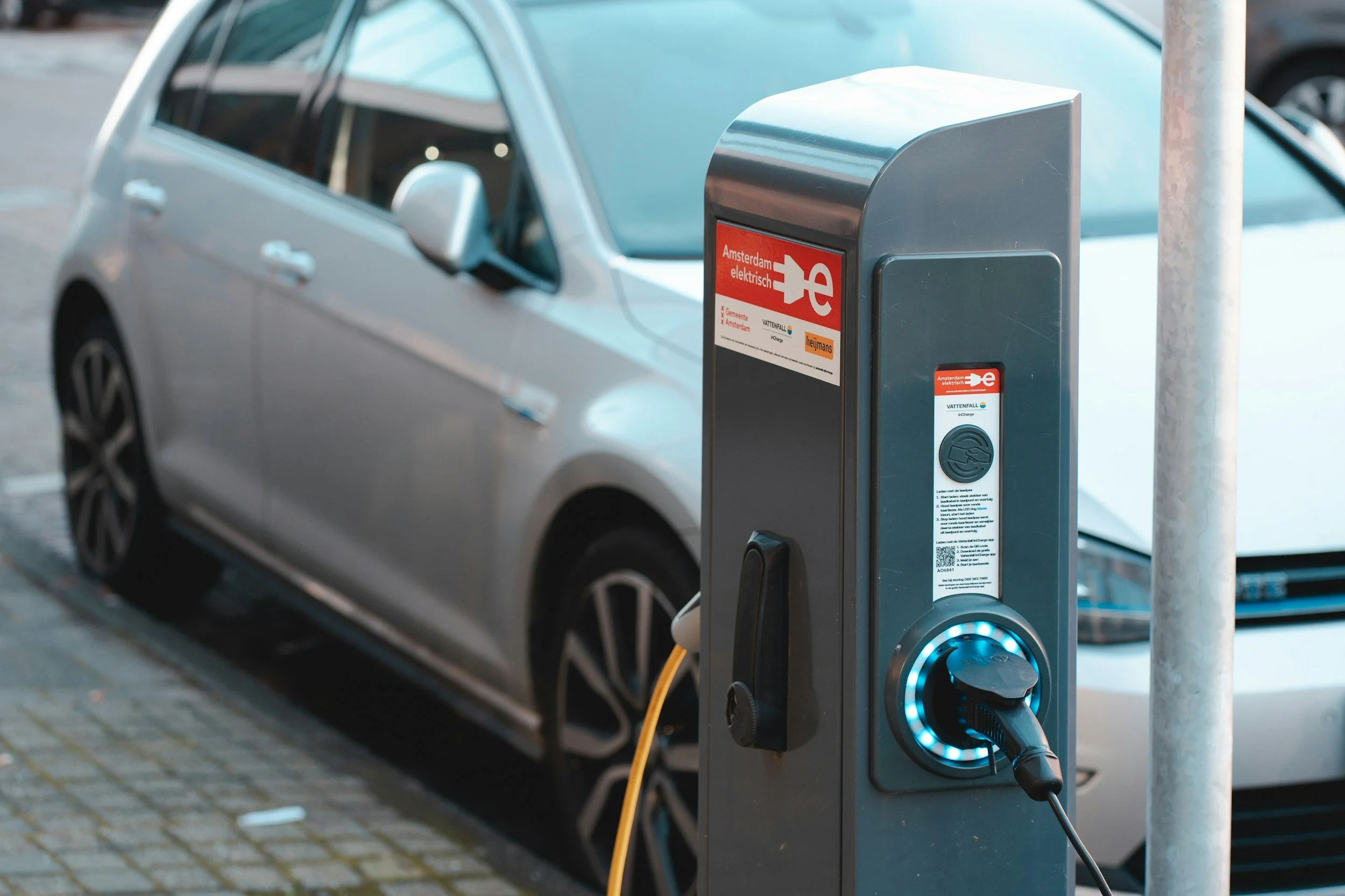 Silver electric car parked at a curbside public charging station pillar along an urban street.