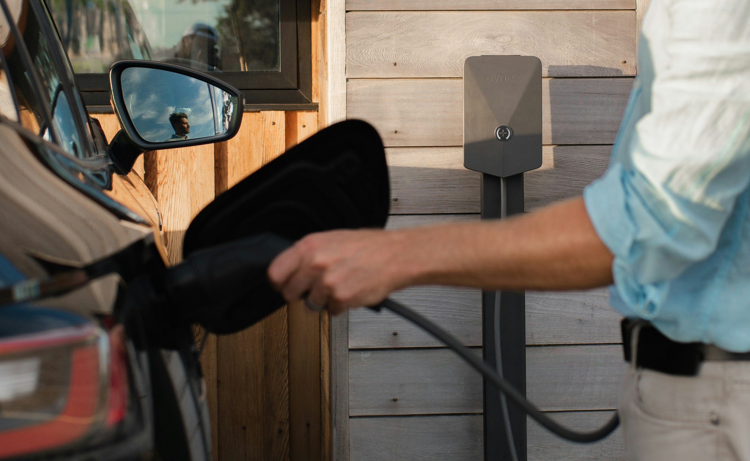 A person plugging a charging cable into an electric car parked next to a wall-mounted charging station.