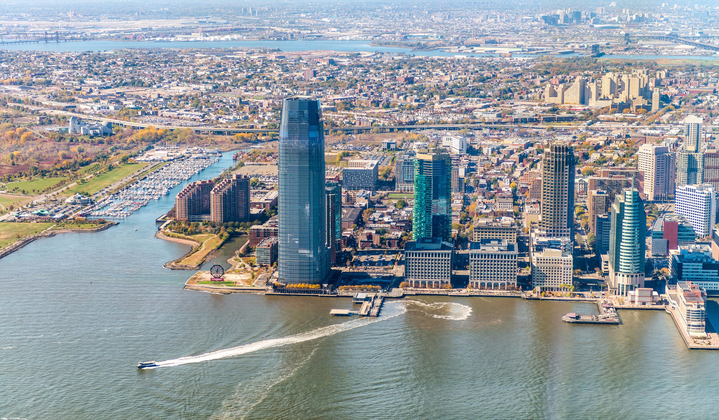 A high-angle aerial view looking across blue water toward modern waterfront condo buildings on a paved river pathway. Behind the buildings is a massive, tree-covered cliff escarpment, topped by a row of diverse houses against a clear blue sky.