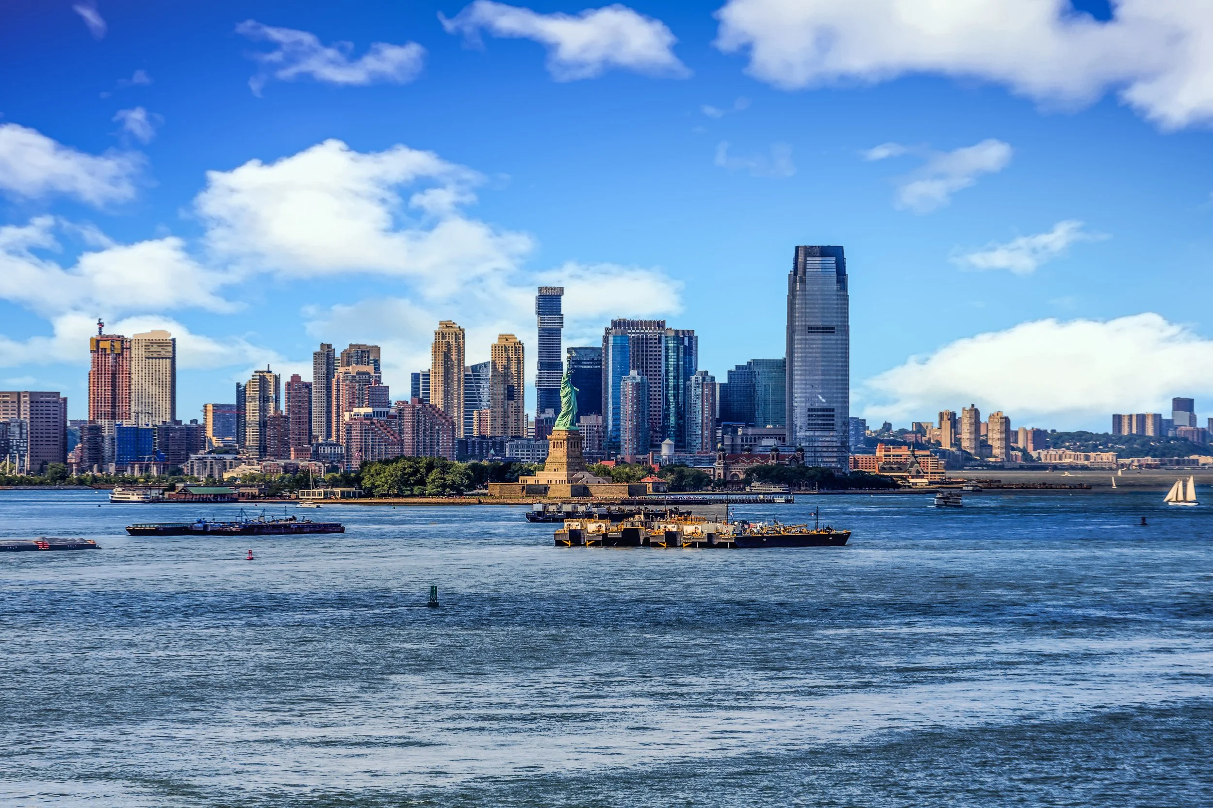 Jersey City skyline and the Statue of Liberty viewed from the water on a sunny day, representing the local Hudson County real estate market (HCMLS / RMLSNJ).