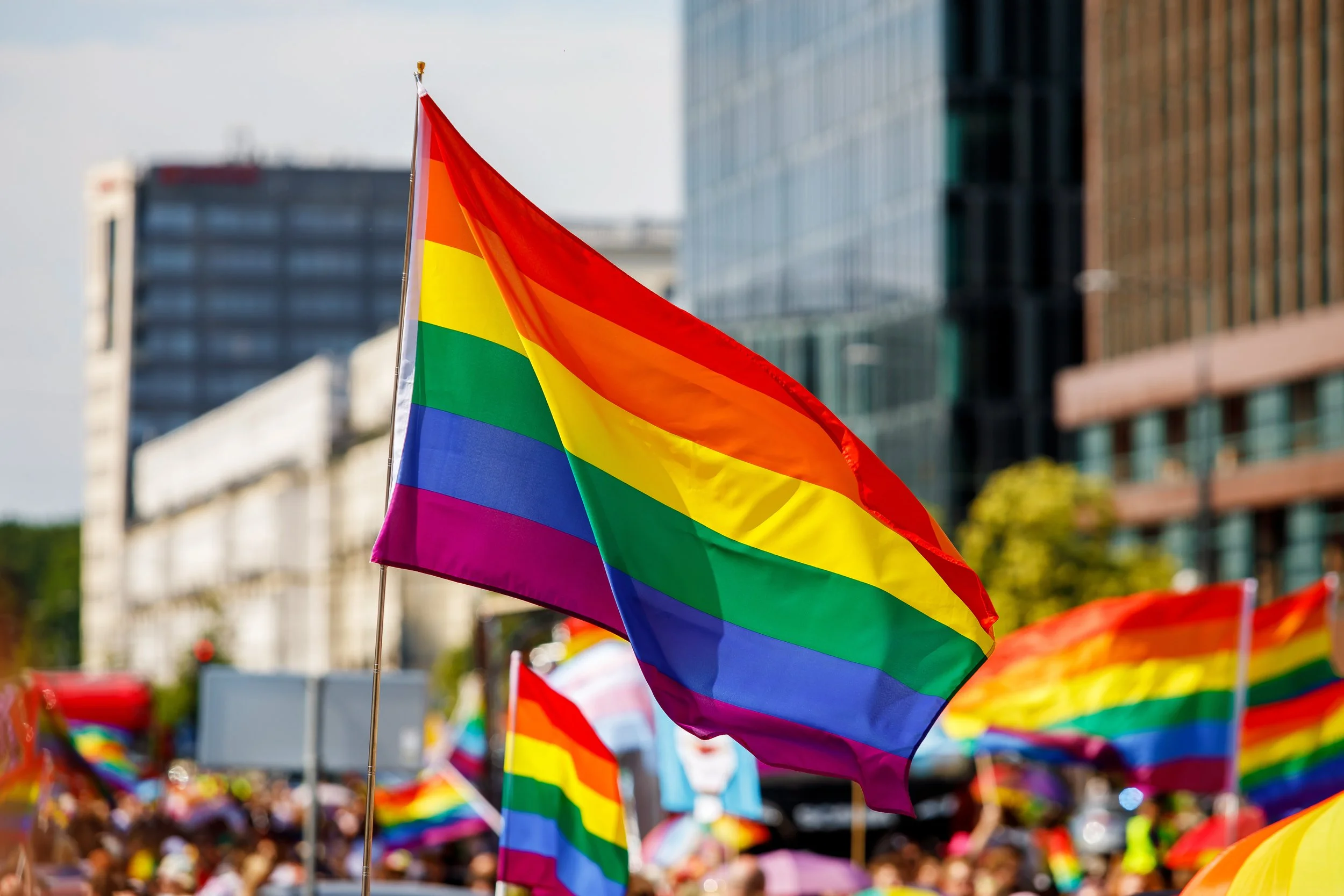 A crisp rainbow Pride flag waving in the wind against a backdrop of blurred city buildings and festival attendees.