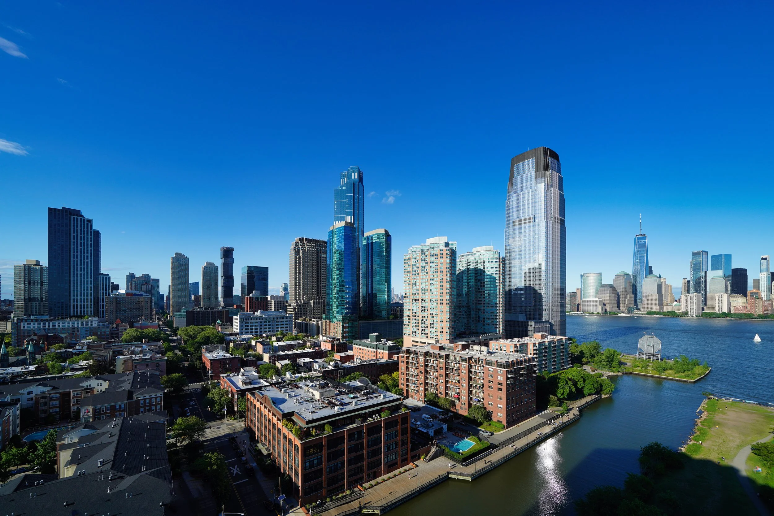 A vibrant, high-angle view of the Jersey City skyline featuring glass skyscrapers like the Goldman Sachs Tower, overlooking the Hudson River and Morris Canal Basin under a clear blue sky.