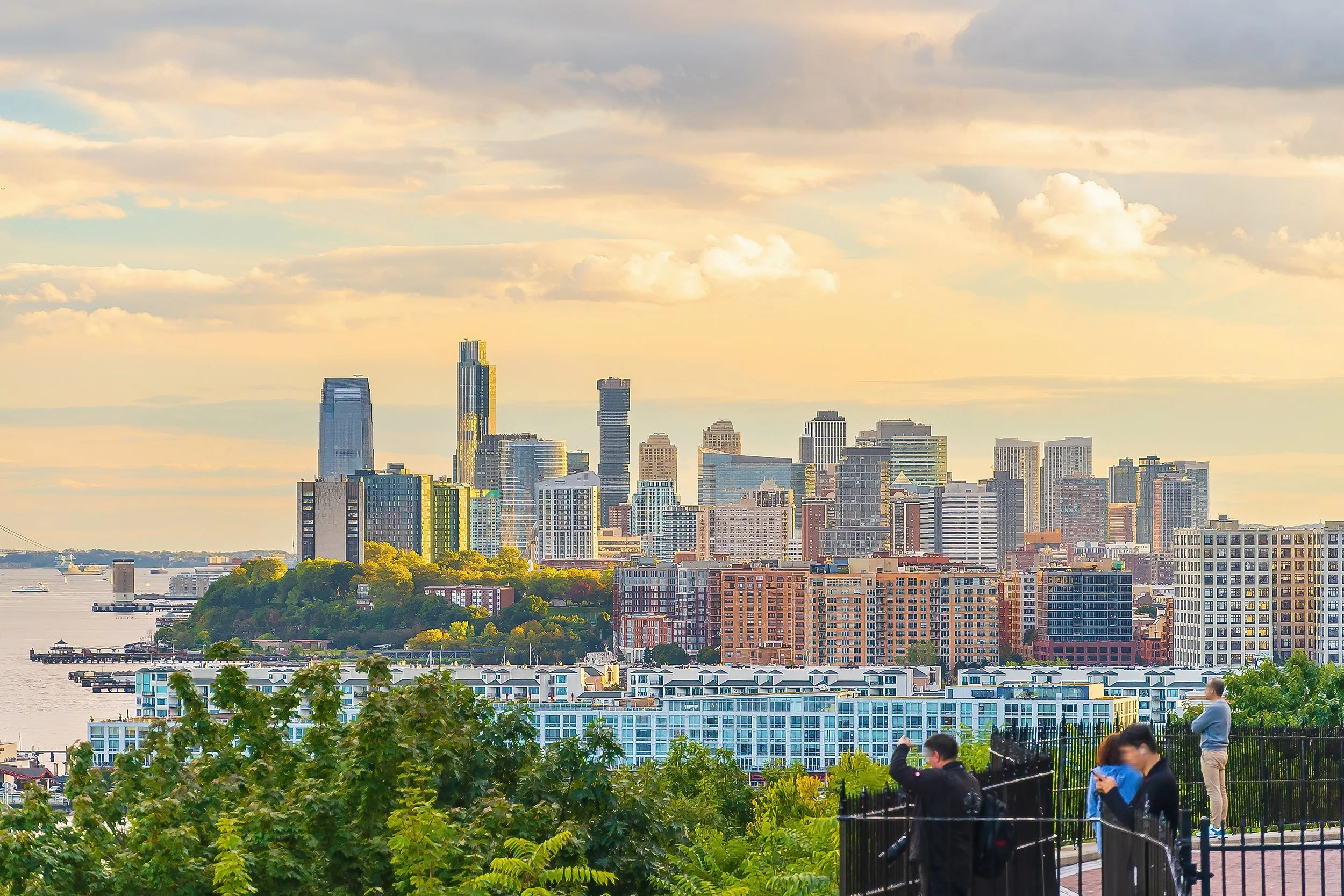 Panoramic view of the Jersey City skyline and Hudson River waterfront during golden hour from an elevated lookout point.