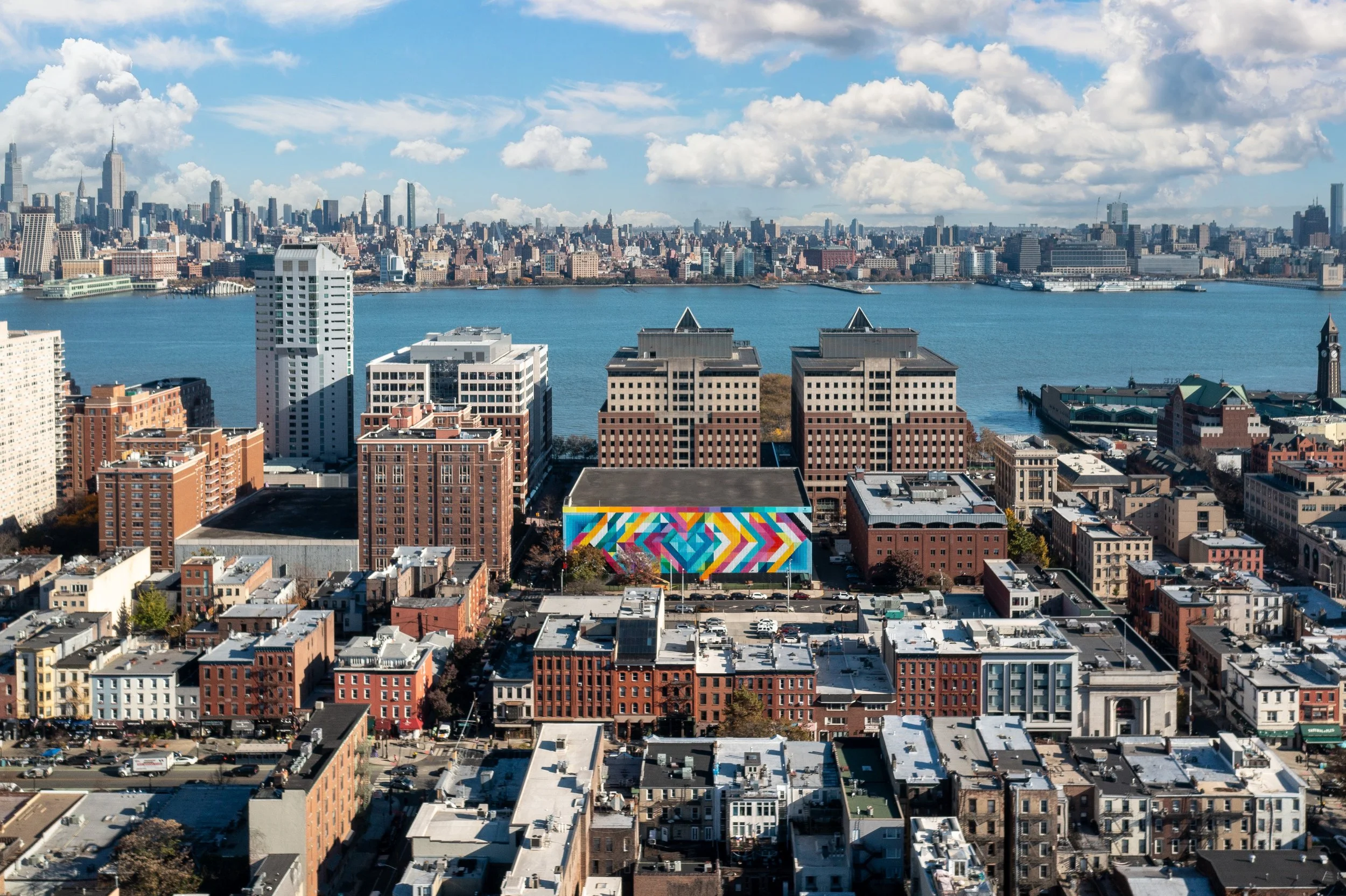 High-angle panoramic view of Jersey City and Hoboken cityscape with the Hudson River and New York City skyline in the background.