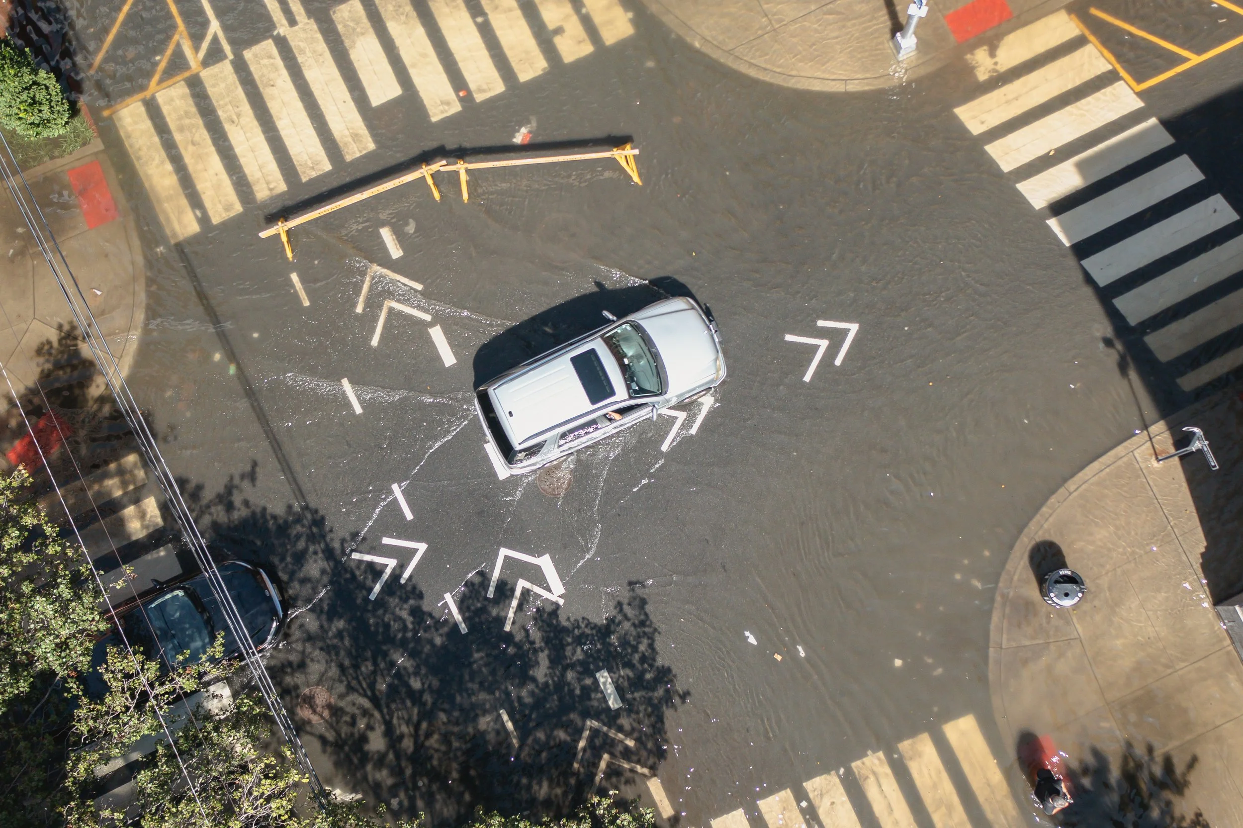 Top-down aerial view of a silver SUV driving through floodwater covering an urban street intersection with crosswalk markings.