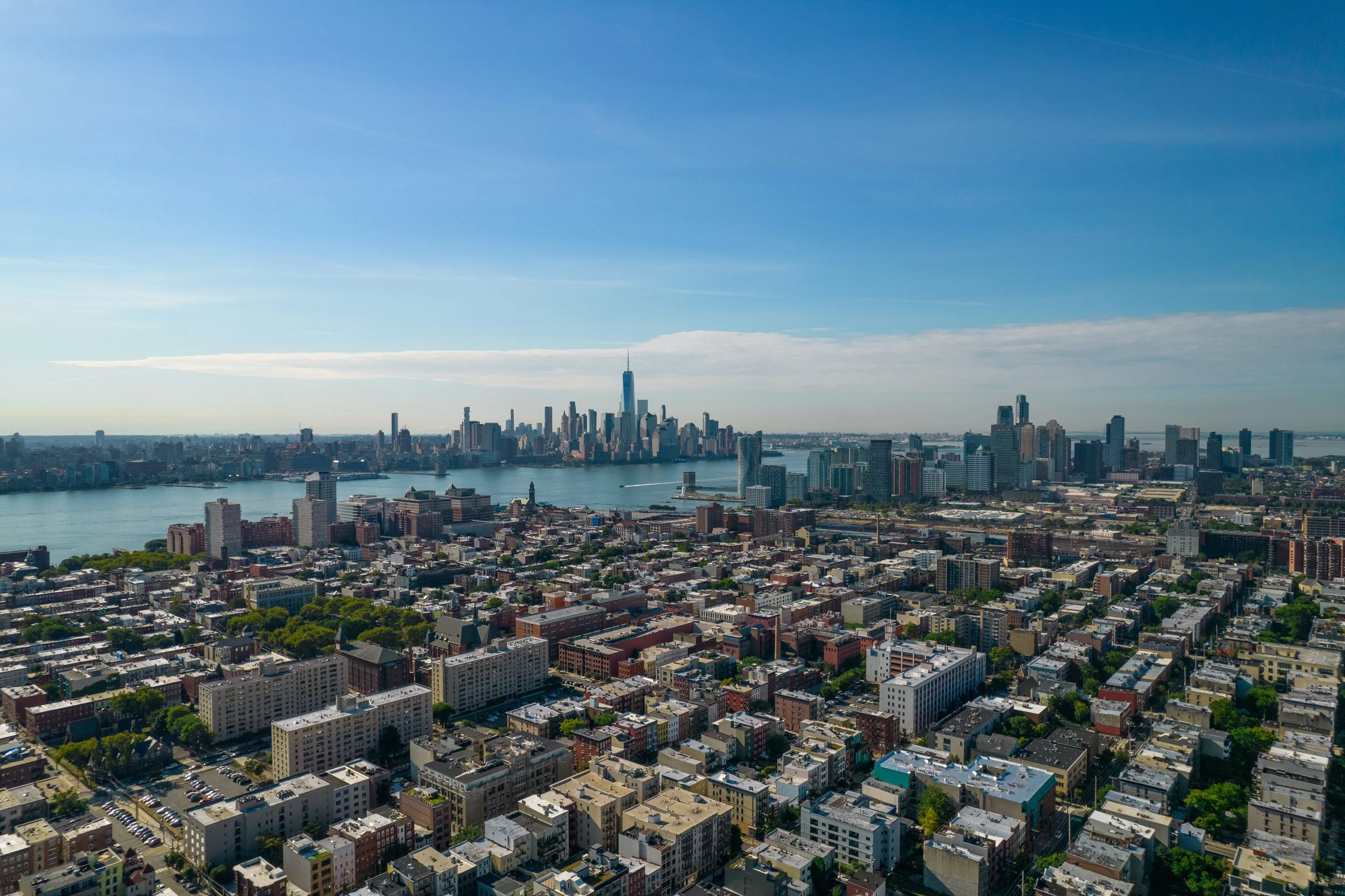 High-angle aerial shot of Hoboken's urban street grid and residential rooftops extending toward the Hudson River and Manhattan skyline.