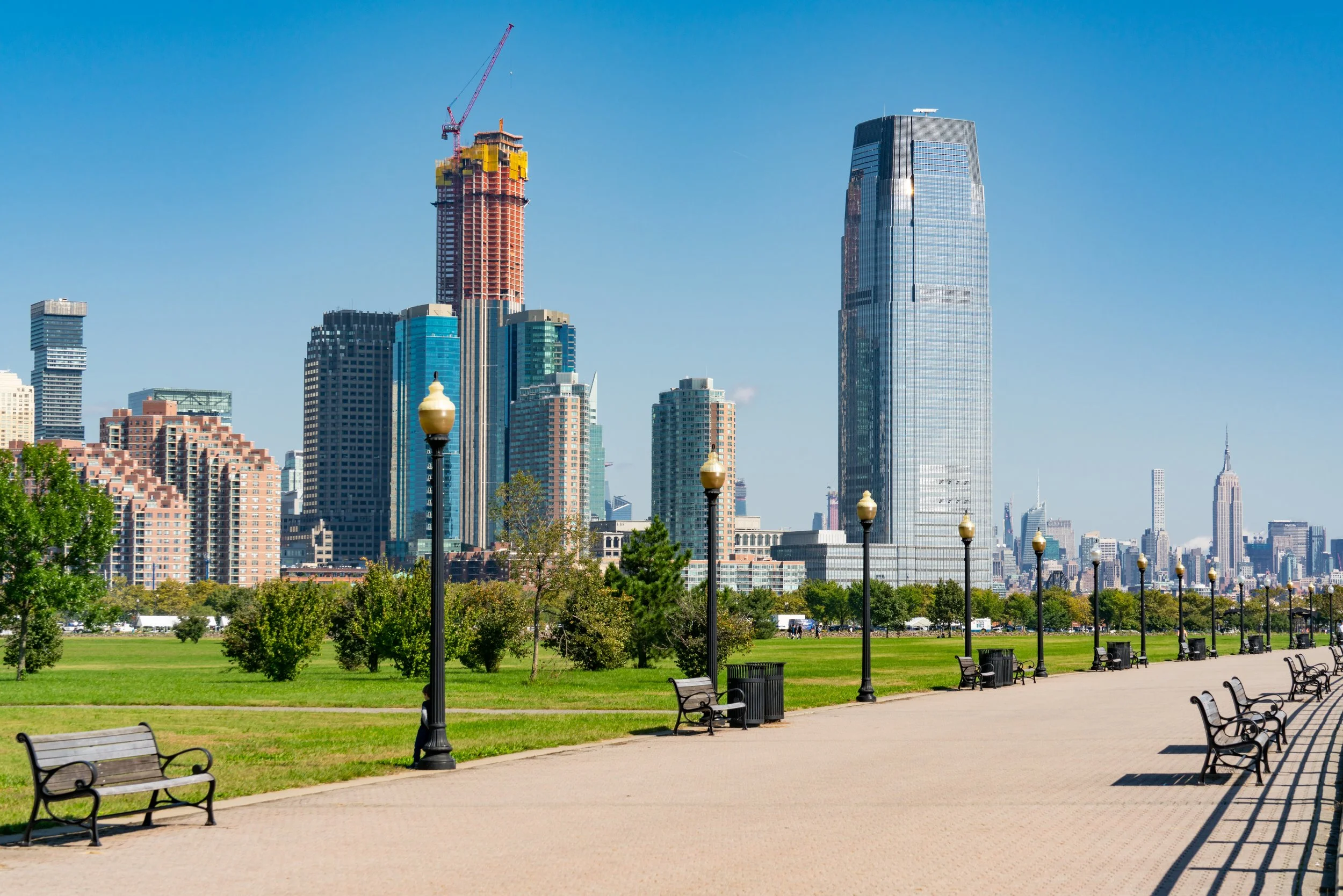 View of the Downtown Jersey City high-rise skyline and waterfront promenade with benches and lamp posts under a clear blue sky.