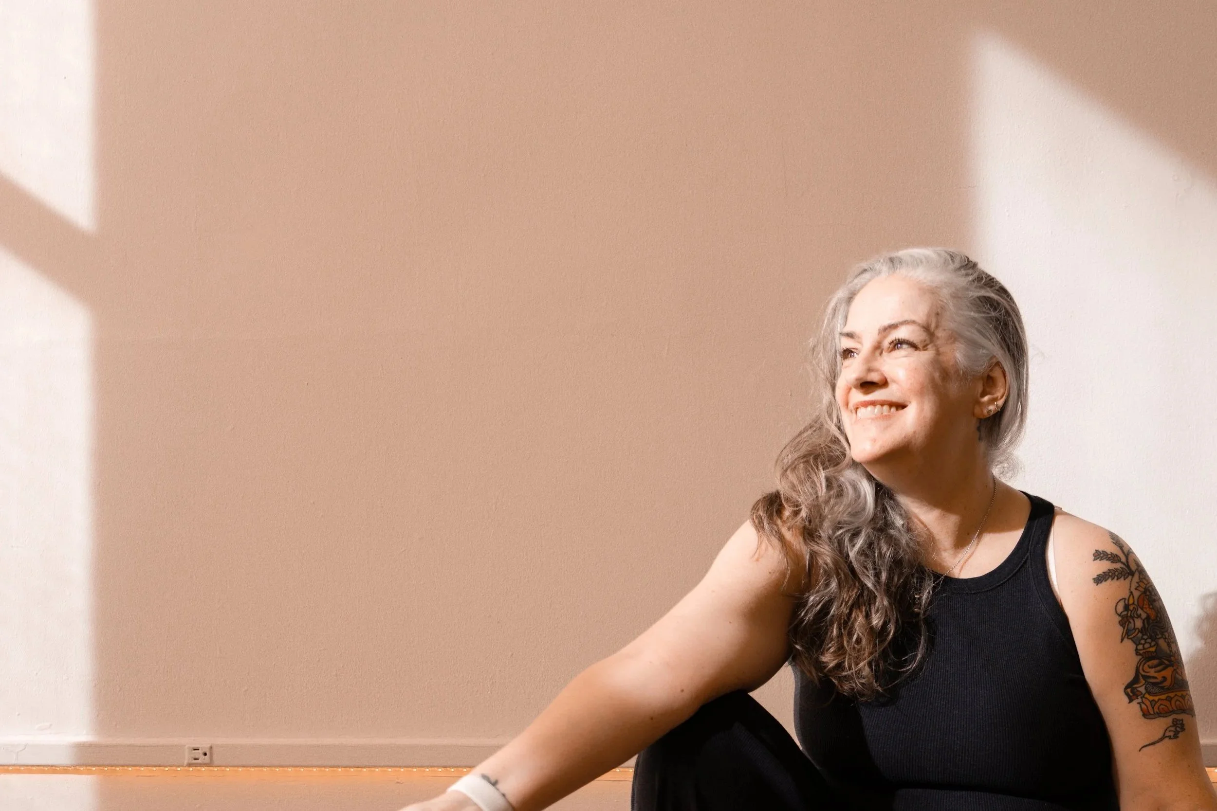 A woman with long gray curly hair, tattoos on her arm, wearing a black sleeveless top, sitting in front of a beige wall with sunlight creating shadows.