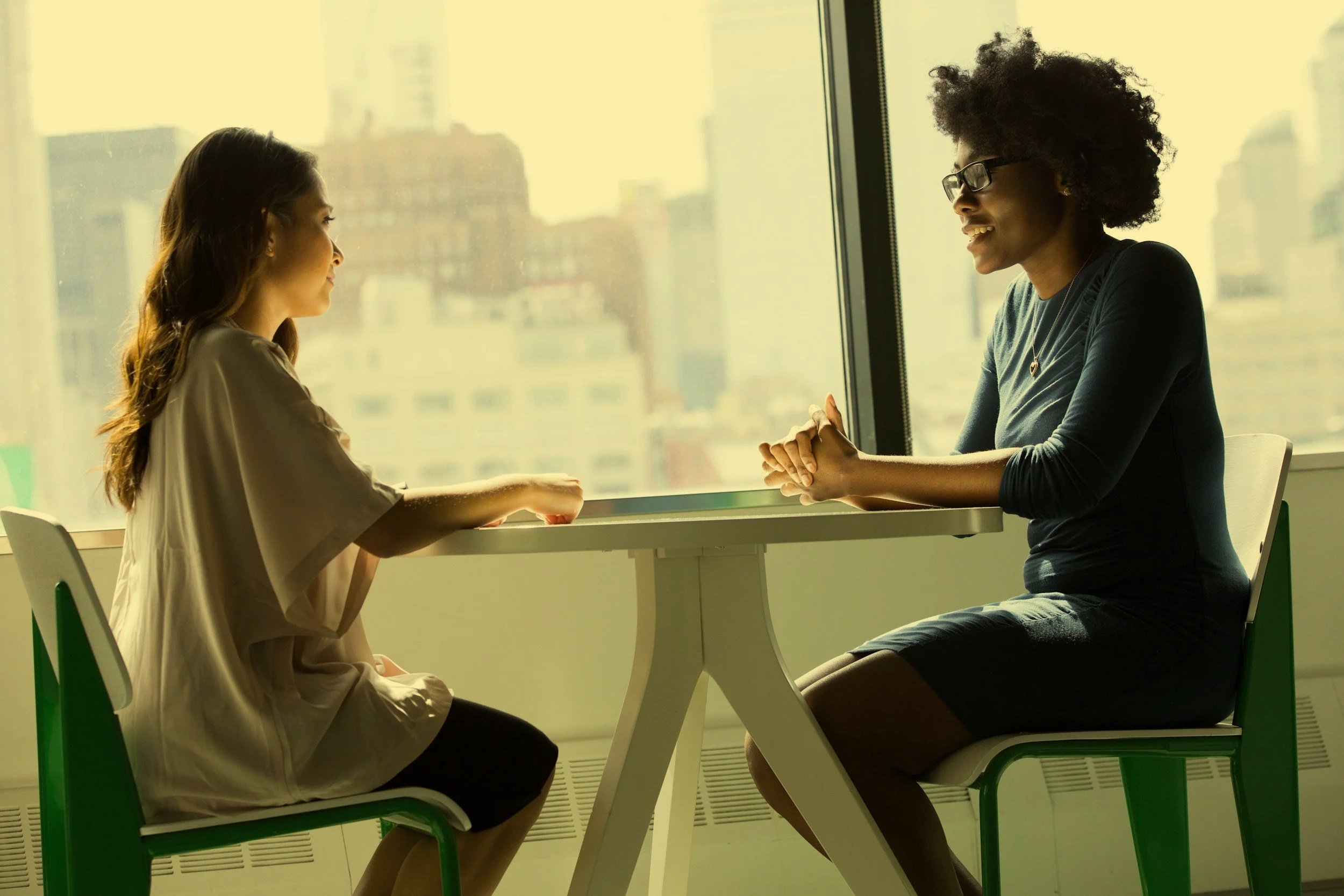 Two women sitting across from each other at a table near a large window, engaging in conversation, with city buildings visible outside.