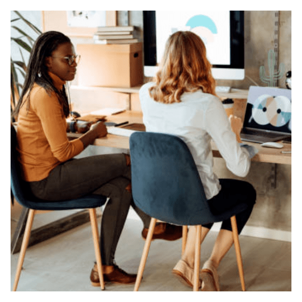 Two women facing away from the viewer working on their computer and talking.