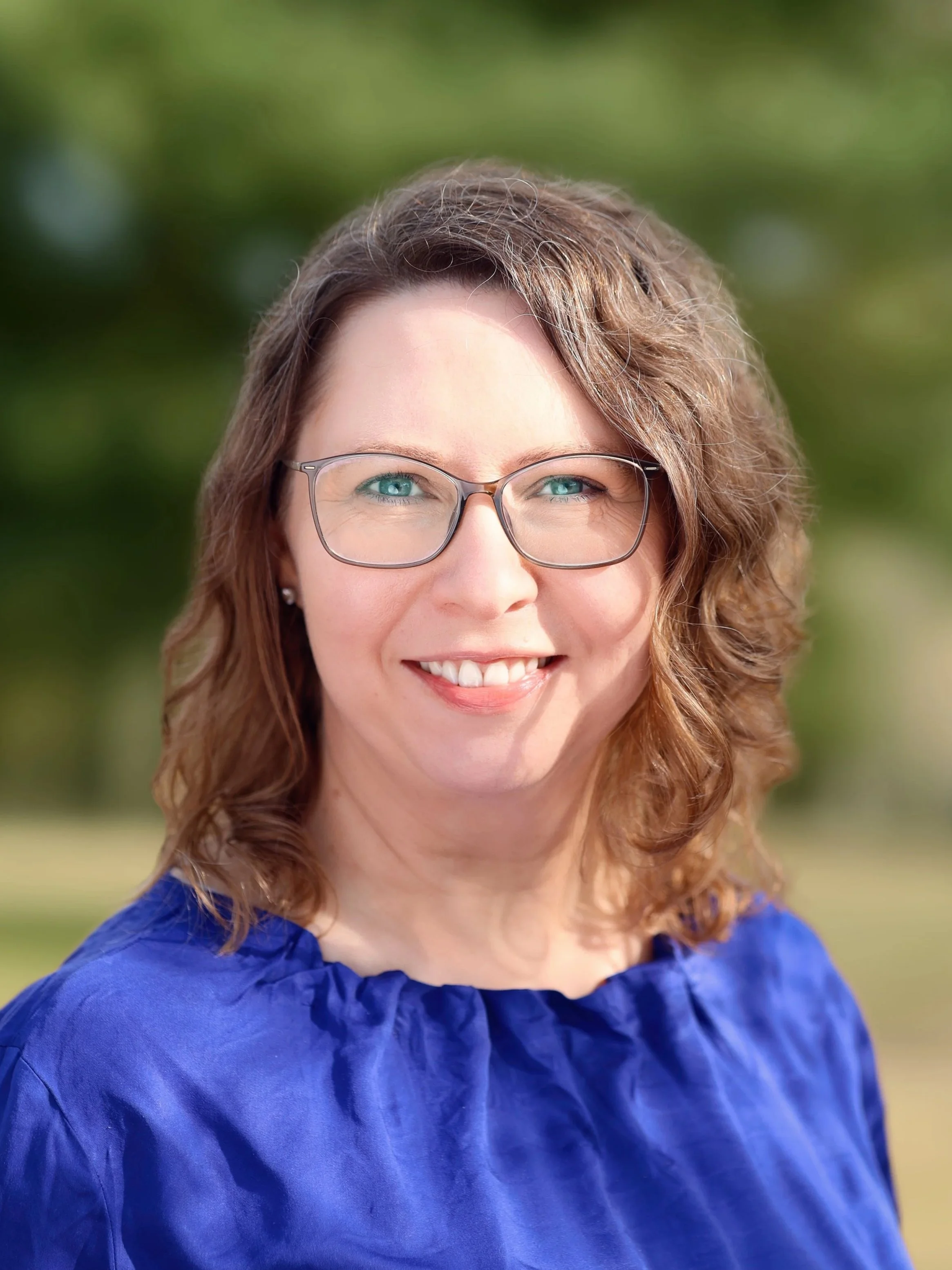 Nahaufnahme einer lächelnden Frau mit braunen, gewellten Haaren, Brille und blauen Oberteil im Freien, im Hintergrund unscharfe grüne Natur.