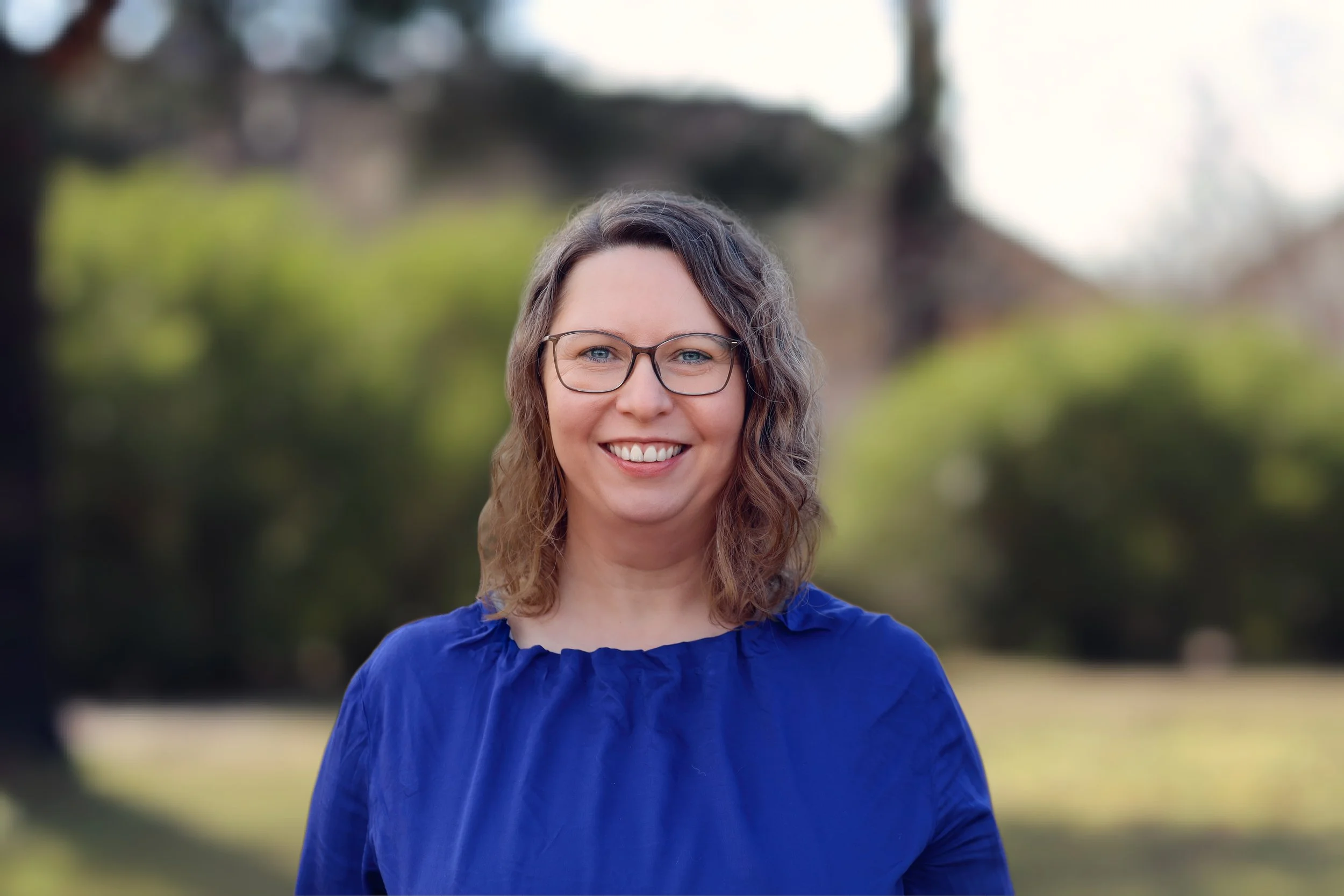 Eine lächelnde Frau mit Brille, braunen Haaren und blauer Bluse, stehend im Park mit grünen Büschen und Bäumen im Hintergrund.