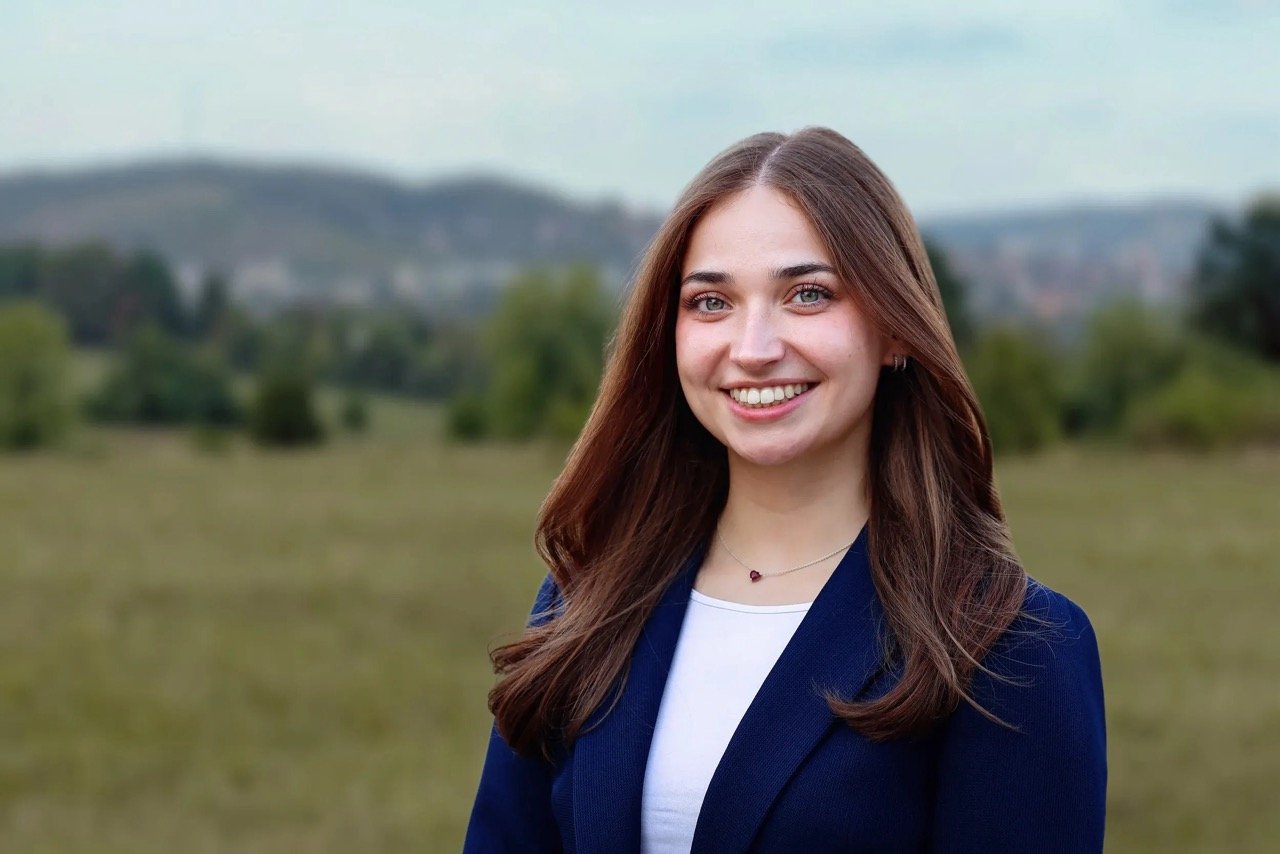 Junge Frau mit braunen Haaren, blauer Blazer und White T-Shirt steht im Freien vor einer grünen Landschaft mit Bäumen und Hügeln im Hintergrund, lächelnd in die Kamera schauend.
