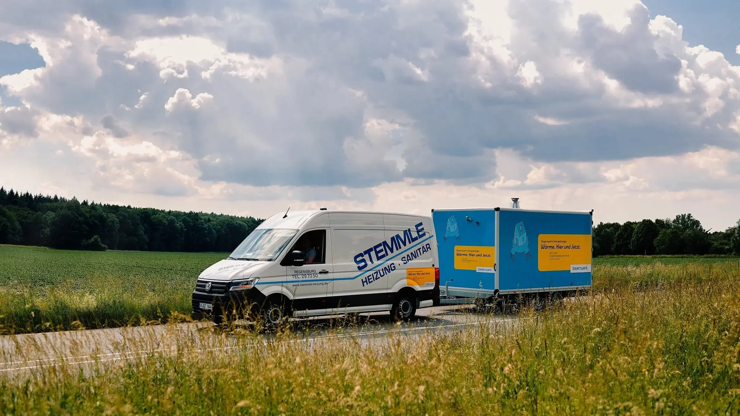 Ein weißer Lieferwagen mit der Aufschrift 'STEMMLE HEIZUNG SANITÄR' fährt auf einer Landstraße, gezogen von einem blauen Anhänger mit einem Roboterlogo und Slogans. Der Himmel ist bewölkt, das Feld ist grün und die Umgebung ist ländlich.