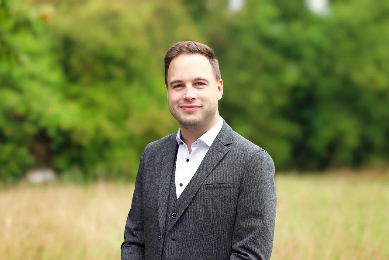 Junge Mann in Anzug auf einer grünen Wiese mit Bäumen im Hintergrund.