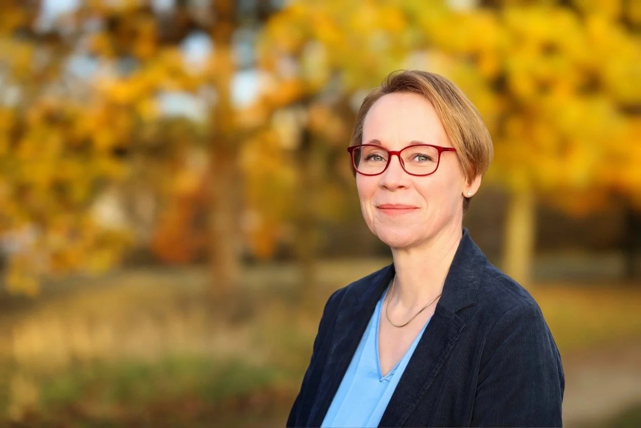 Porträt einer Frau mit roten Brillen, kurzen braunen Haaren, dunklem Jackett und blauer Bluse vor herbstlichem Hintergrund mit gelben Blättern.