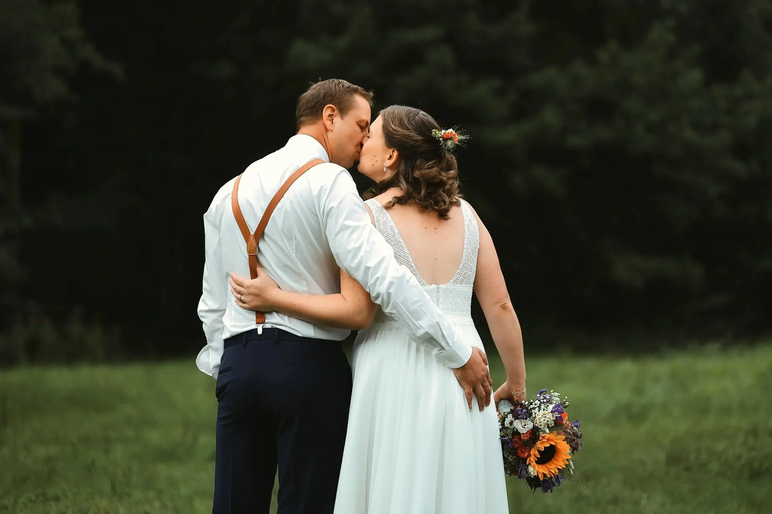 Ein verliebtes Brautpaar bei einem Kuss im Grünen, die Frau hält einen Blumenstrauß, die Szene ist romantisch und natürlich.