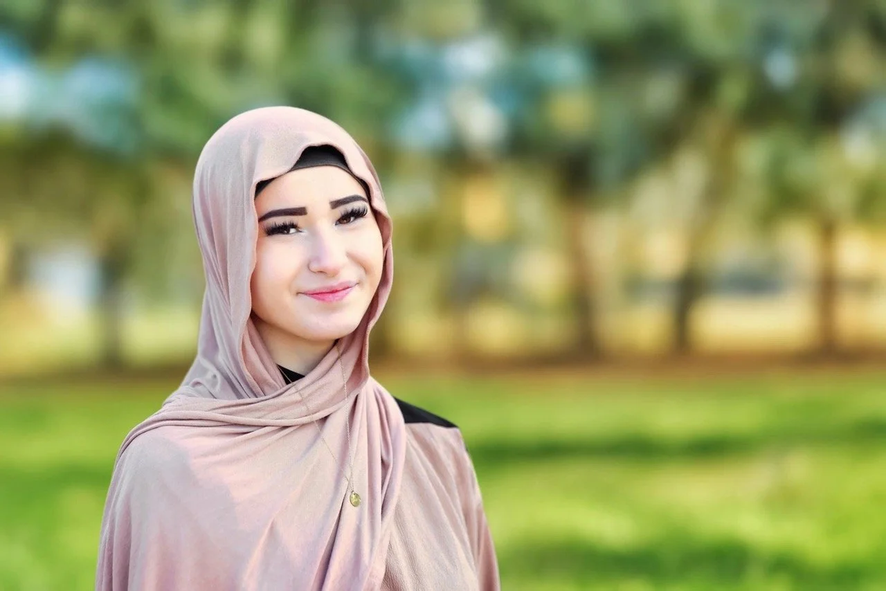 Junge Frau mit schwarzem Kopftuch und beigefarbenem Hijab in einem Park auf grünem Rasen mit Bäumen im Hintergrund, lächelnd.