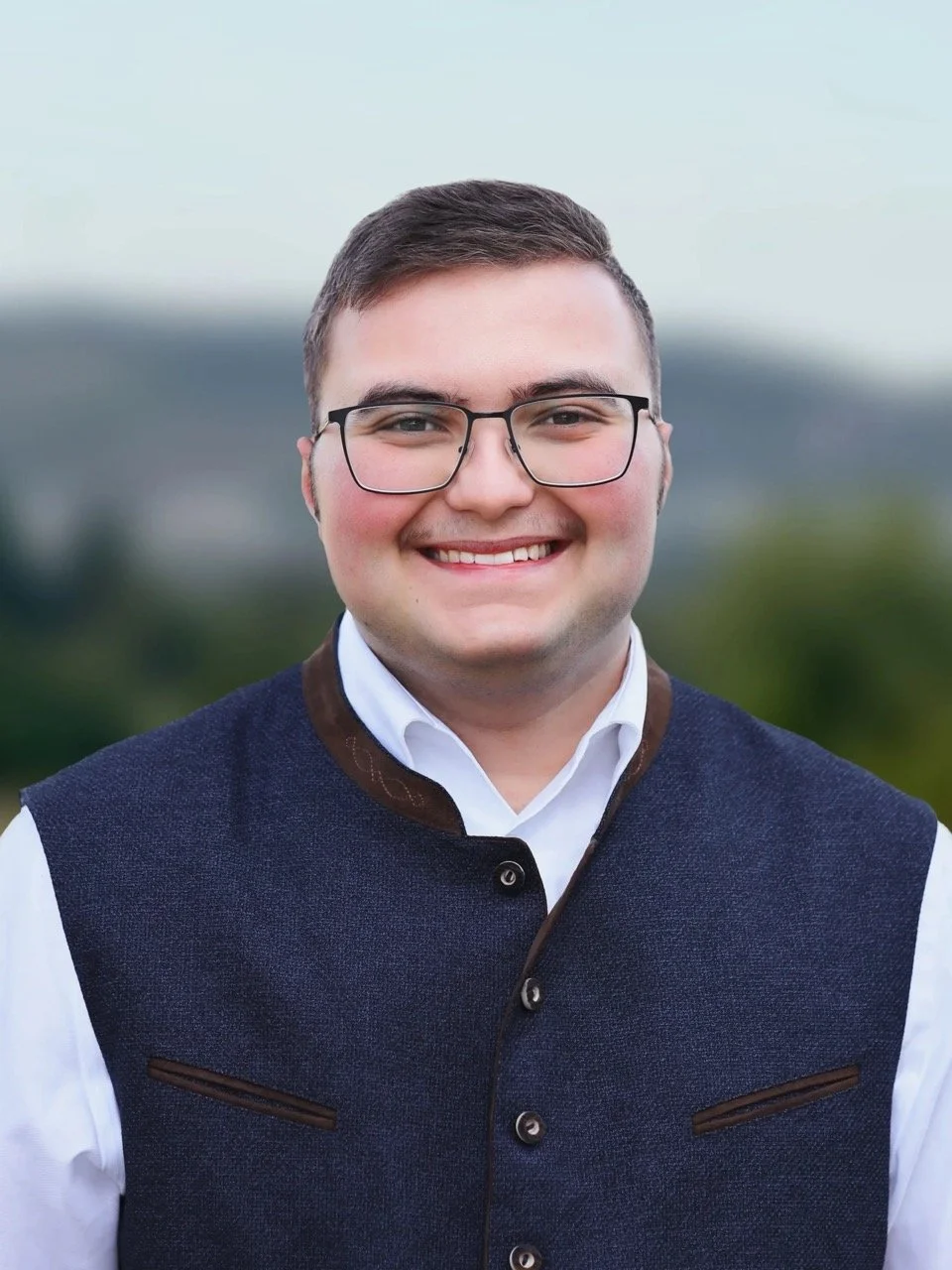 Porträt eines lächelnden jungen Mannes mit Brille, dunklen Haaren, weißem Hemd und dunkelblauer Weste, im Freien mit unscharfem grünen und blauen Hintergrund.