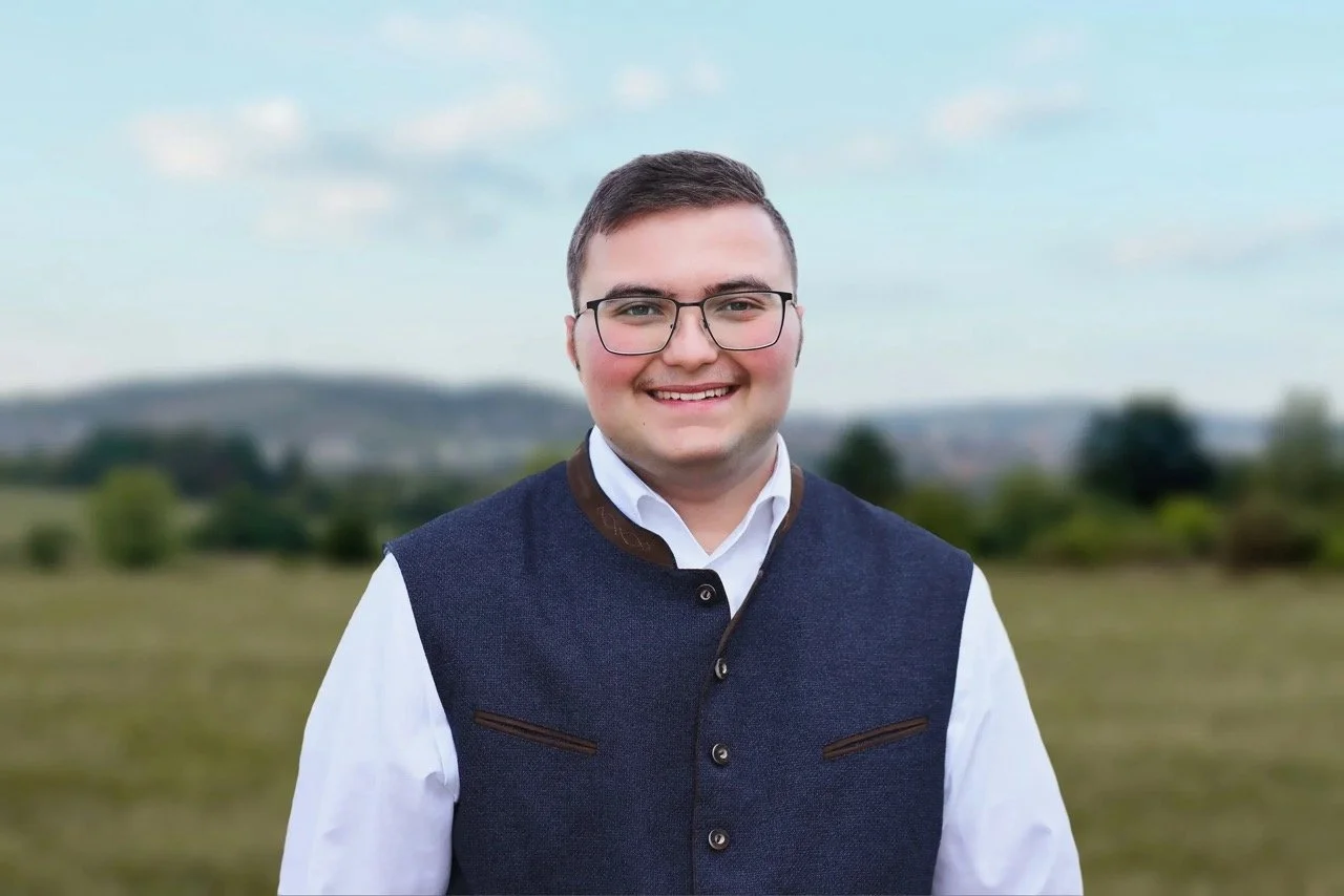Ein junger Mann mit Brille und traditioneller Trachtenweste, lächelnd, im Freien mit Hügeln und Bäumen im Hintergrund.