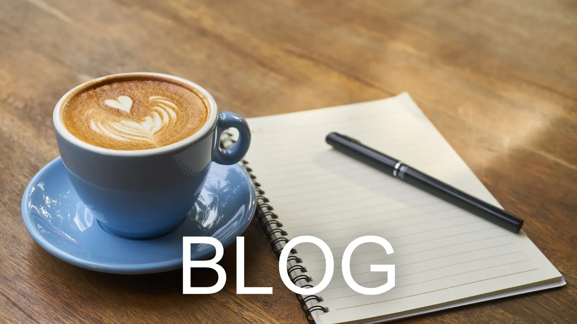 Latte Art in einer blauen Tasse neben Notizbuch und Stift auf Holztisch mit "BLOG" Text.