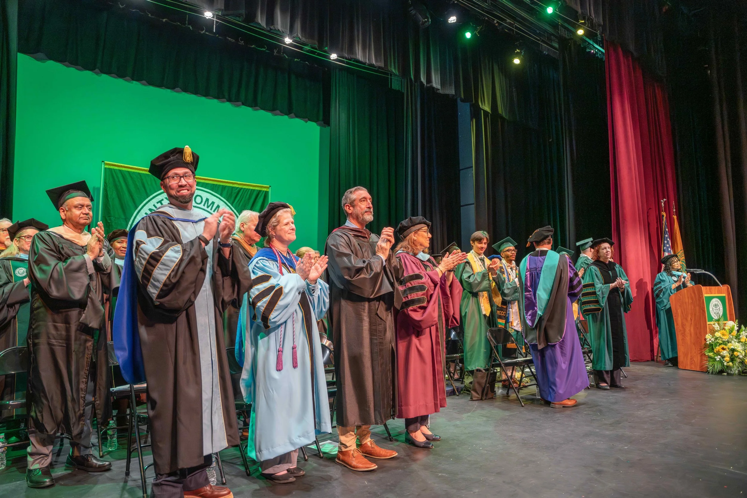 Group of people in academic robes and caps standing on stage during a graduation ceremony, clapping.  College Graduation photography. 