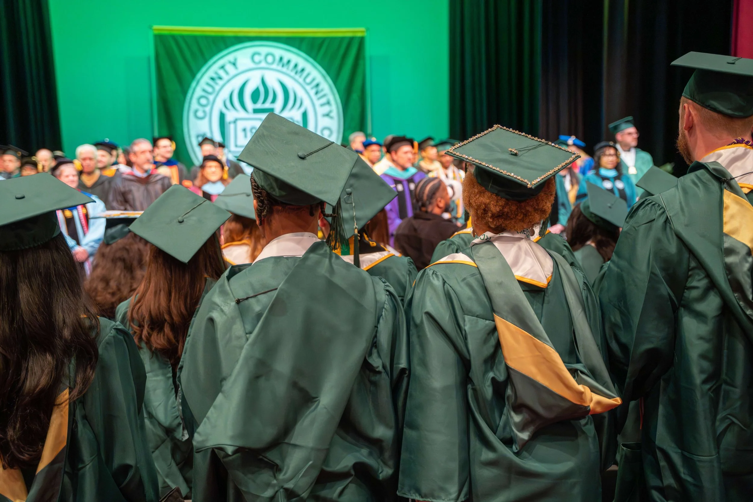 Graduates in green caps and gowns attending a commencement ceremony with a large green banner that reads 'County Community College' in the background. College Graduation photography. 
