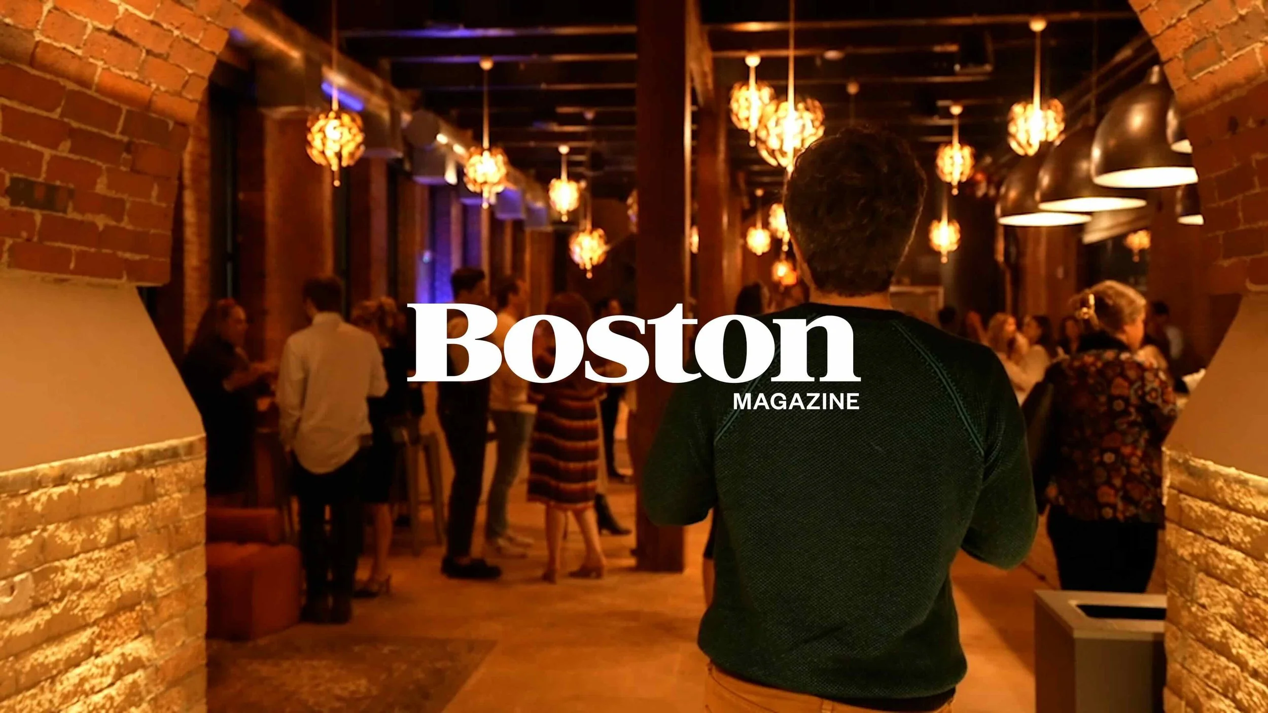 A man with curly hair wearing a black shirt faces a gathering of people in a warmly lit bar or restaurant with exposed brick walls and hanging pendant lights. The text 'Boston magazine' is overlayed on the image.
