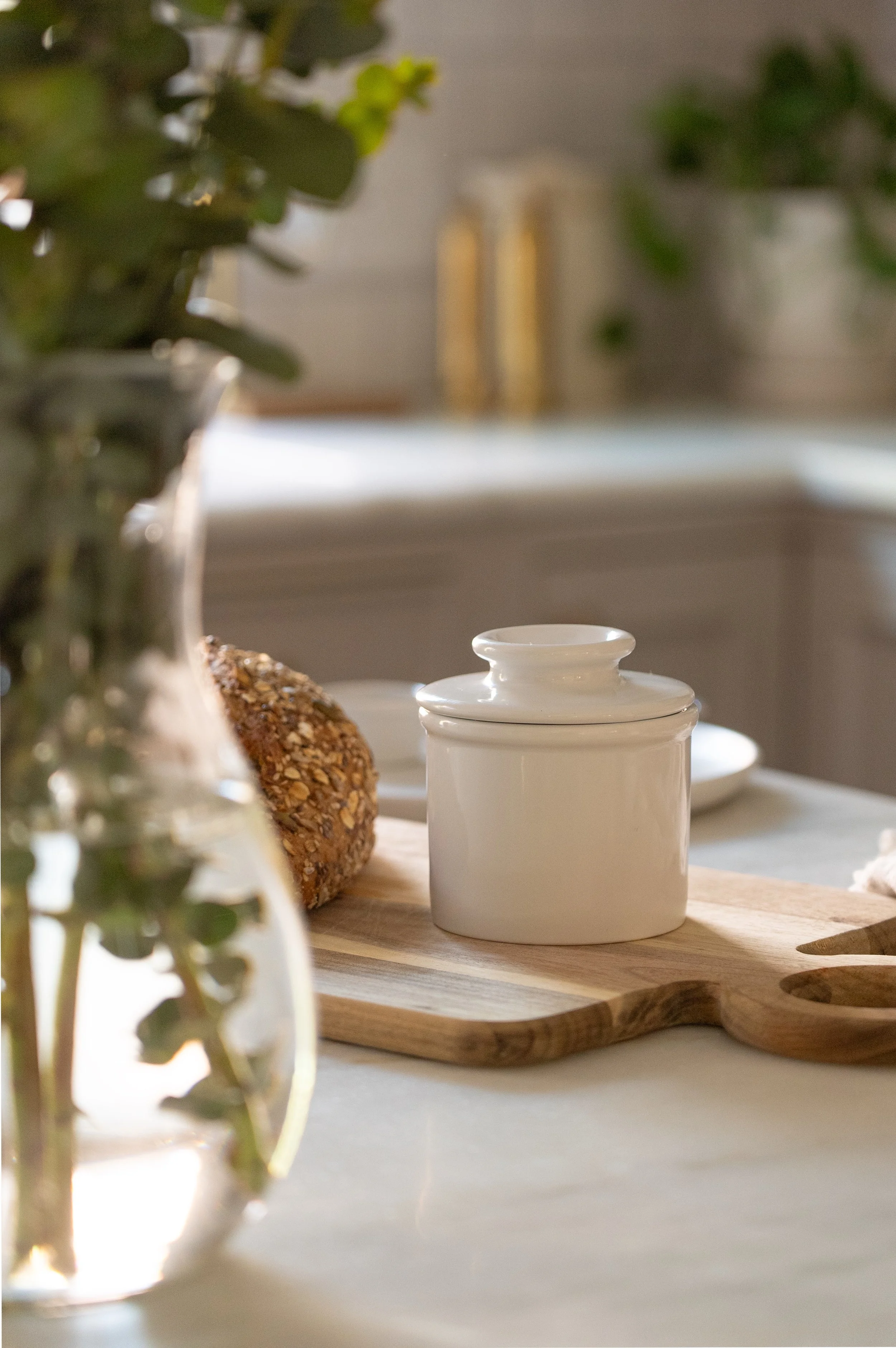 White ceramic jar with lid on a wooden cutting board, next to a loaf of multigrain bread, on a white kitchen countertop with blurred background.