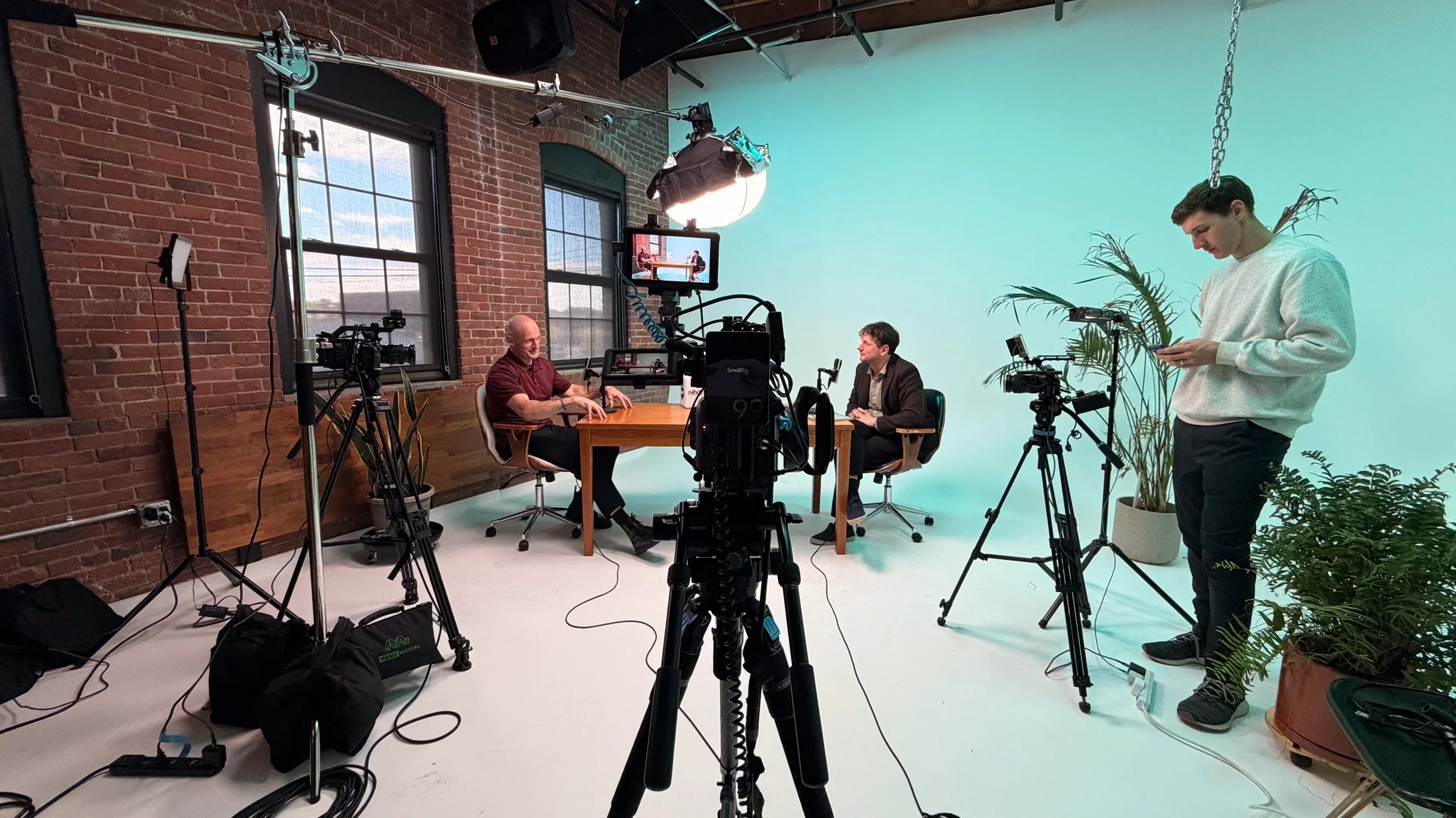 Two men seated at a table in a video recording studio with brick walls and large windows, surrounded by camera equipment, lighting, and plants, with a crew member adjusting his phone.